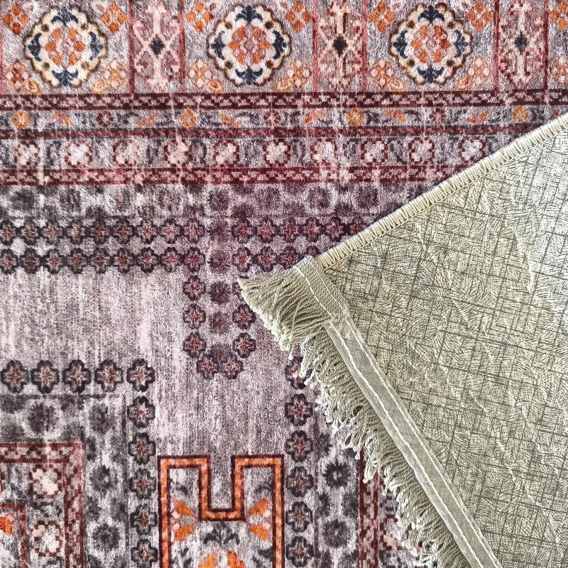 Tapis aux motifs traditionnels gris, orange et bordeaux. Son coin est relevé, révélant le dessous clair quadrillé avec des franges