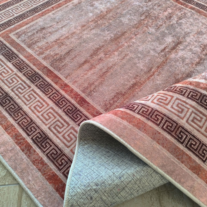 Tapis aux tons terre et marron-rouge, avec un motif central abstrait délavé et une bordure géométrique de méandres, partiellement enroulé