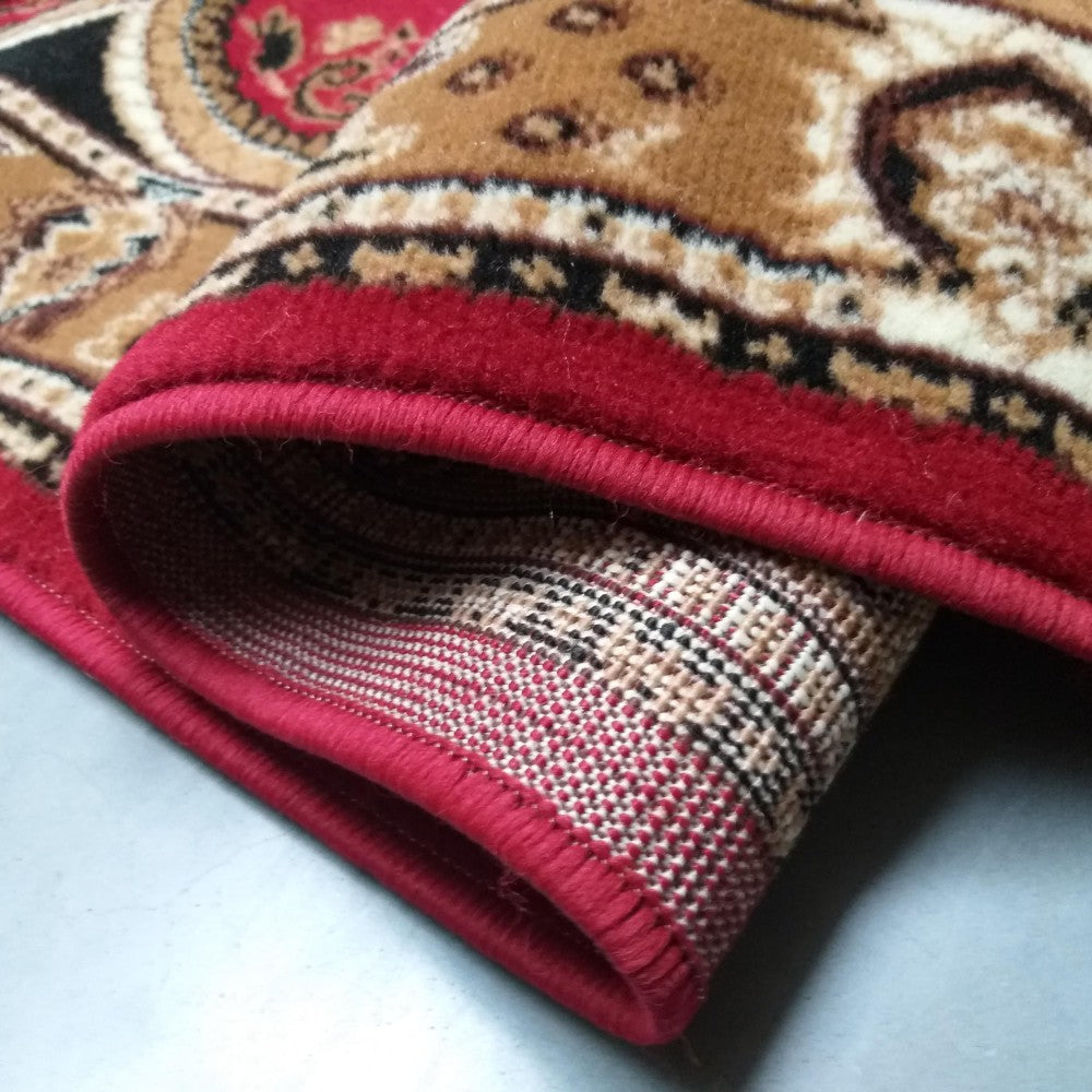 Tapis enroulé avec des motifs traditionnels rouges, bruns et noirs, bordé de rouge. L'envers du tapis révèle une texture tissée