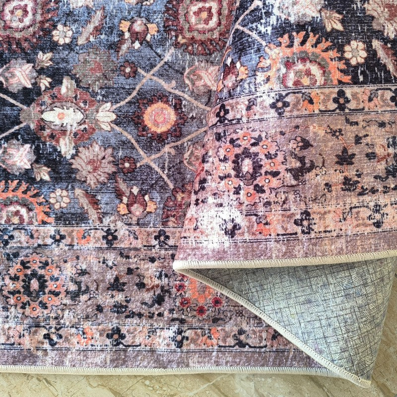 Tapis vintage à motifs floraux complexes et médaillons, teintes bleu-gris, rouille et crème. Un côté est roulé, dévoilant son envers quadrillé clair