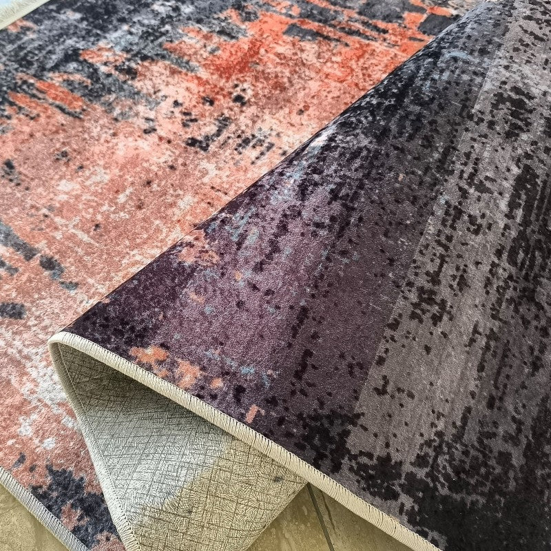 Tapis doux au motif abstrait mêlant le terre cuite, le gris et le noir. Partiellement replié, il révèle son envers clair et son bord surpiqué