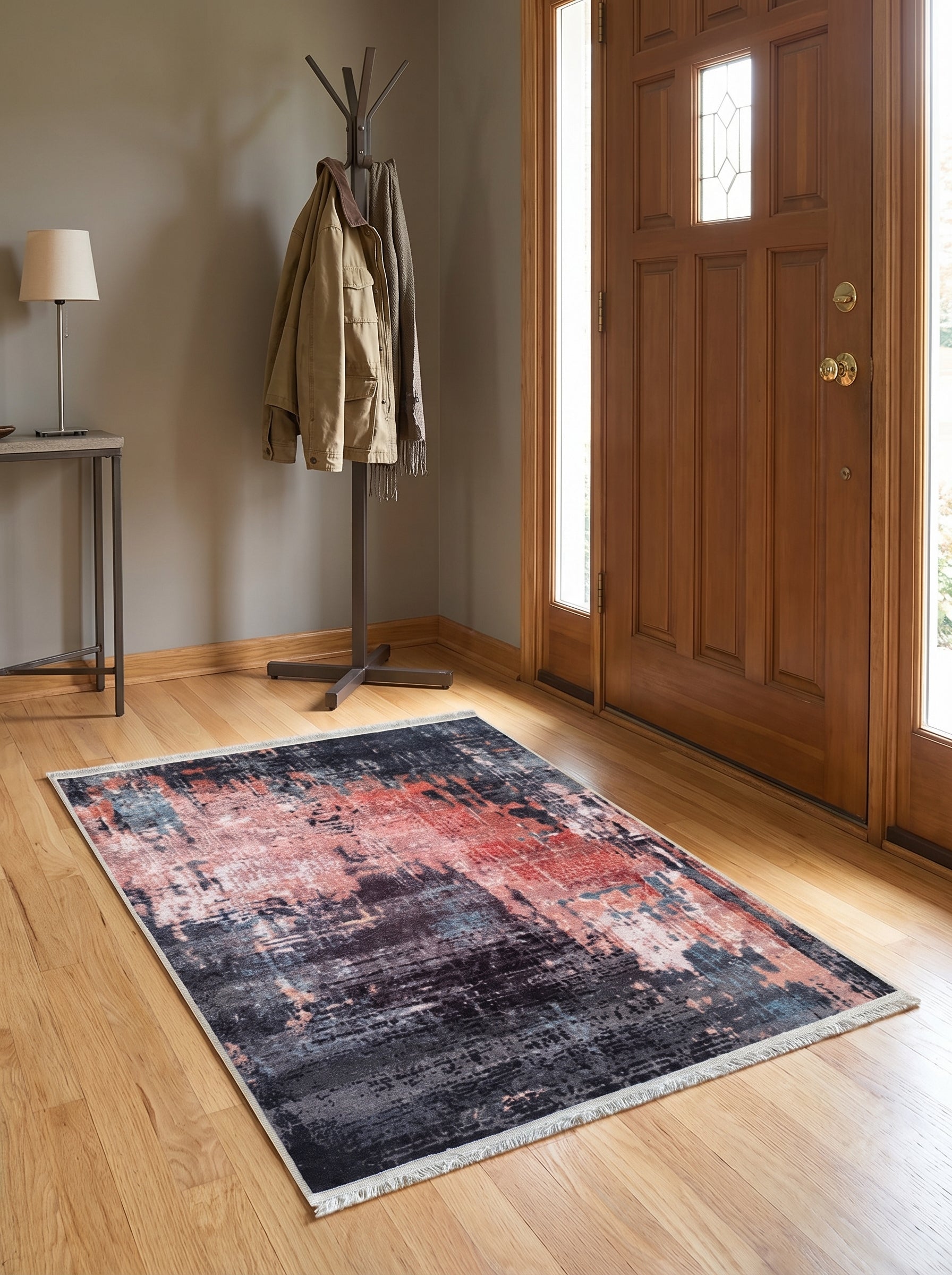 Tapis rectangulaire aux motifs abstraits mêlant rouge corail, gris charbon, noir et bleu, avec des franges claires dans une entrée