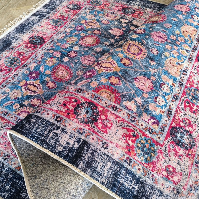 Tapis aux motifs floraux et orientaux colorés en bleu, rose, rouge et or, partiellement roulé, montrant ses bordures sombres et franges blanches
