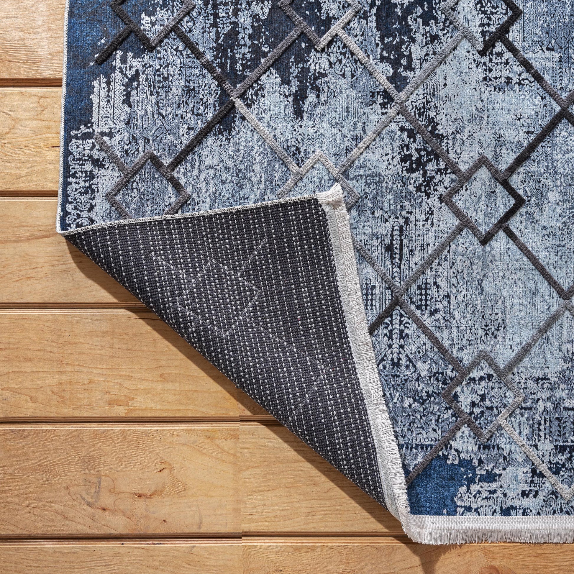 Tapis bleu et blanc à motif abstrait et losanges gris, dont le coin est relevé sur un parquet en bois, montrant le dessous à picots