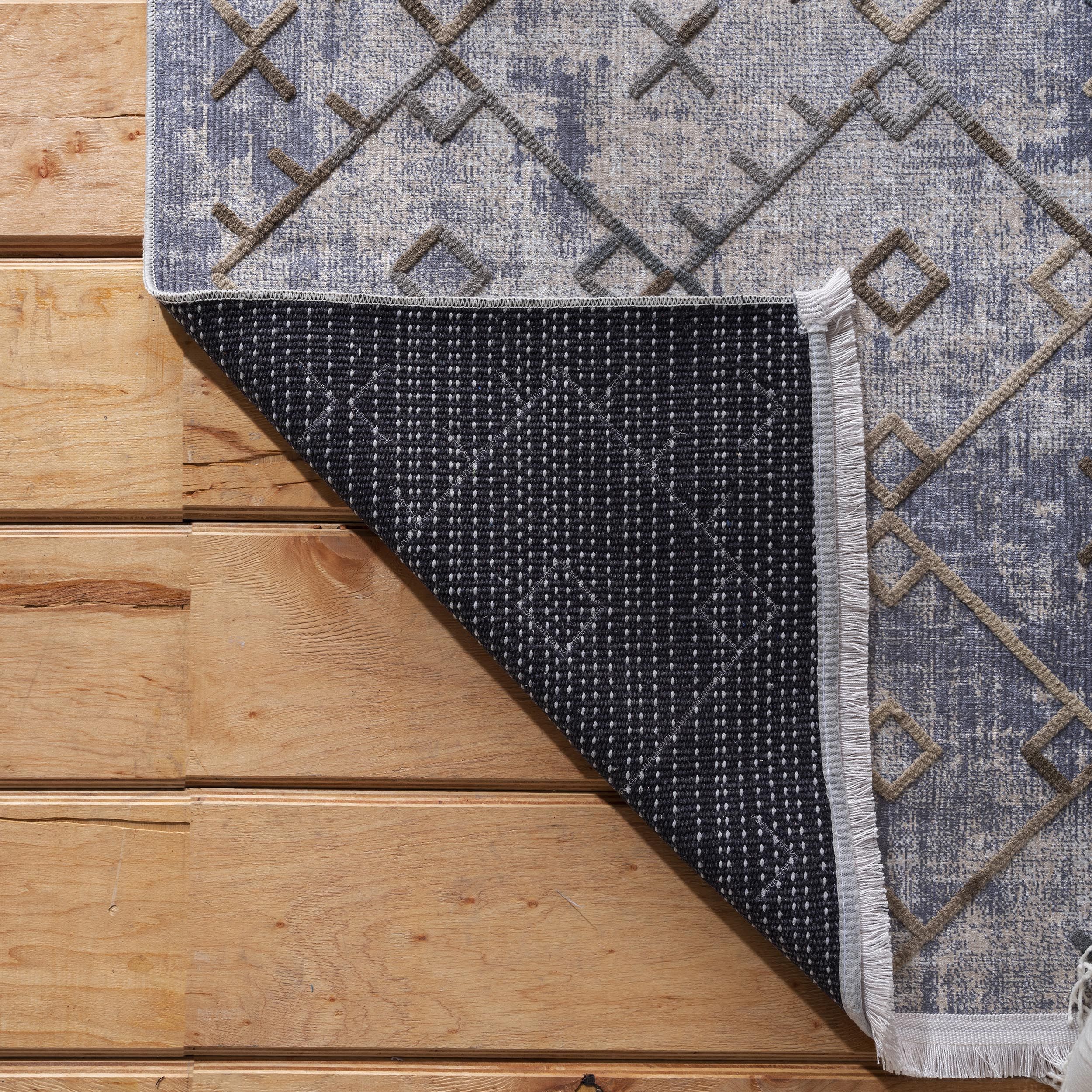 Tapis gris bleu et beige aux motifs géométriques marron en relief, partiellement replié sur un plancher en bois. L'envers noir à petits points blancs et une frange latérale sont visibles