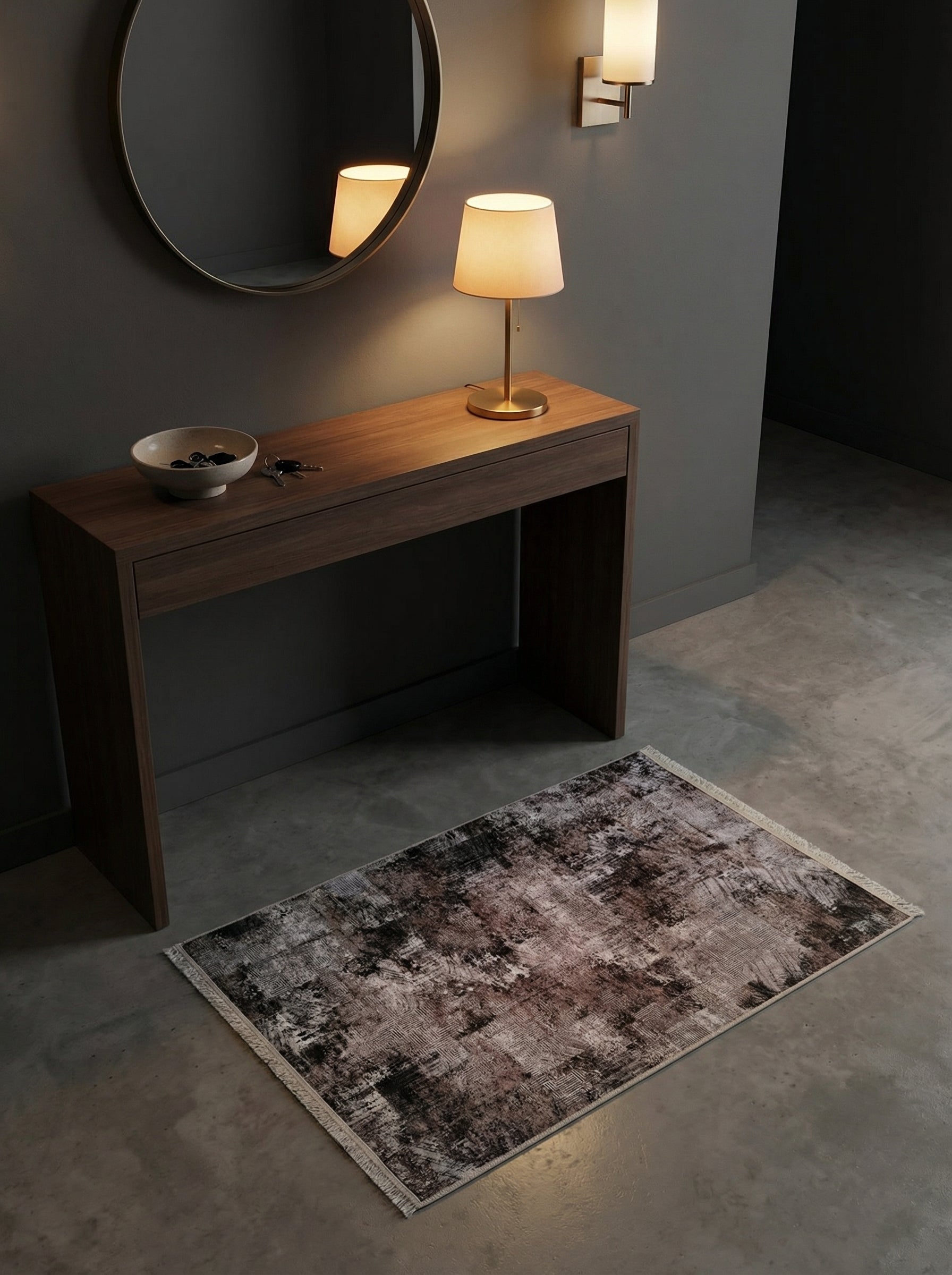 Tapis rectangulaire aux motifs abstraits gris foncé, noirs et bruns délavés avec franges claires, posé dans une entrée au sol en béton