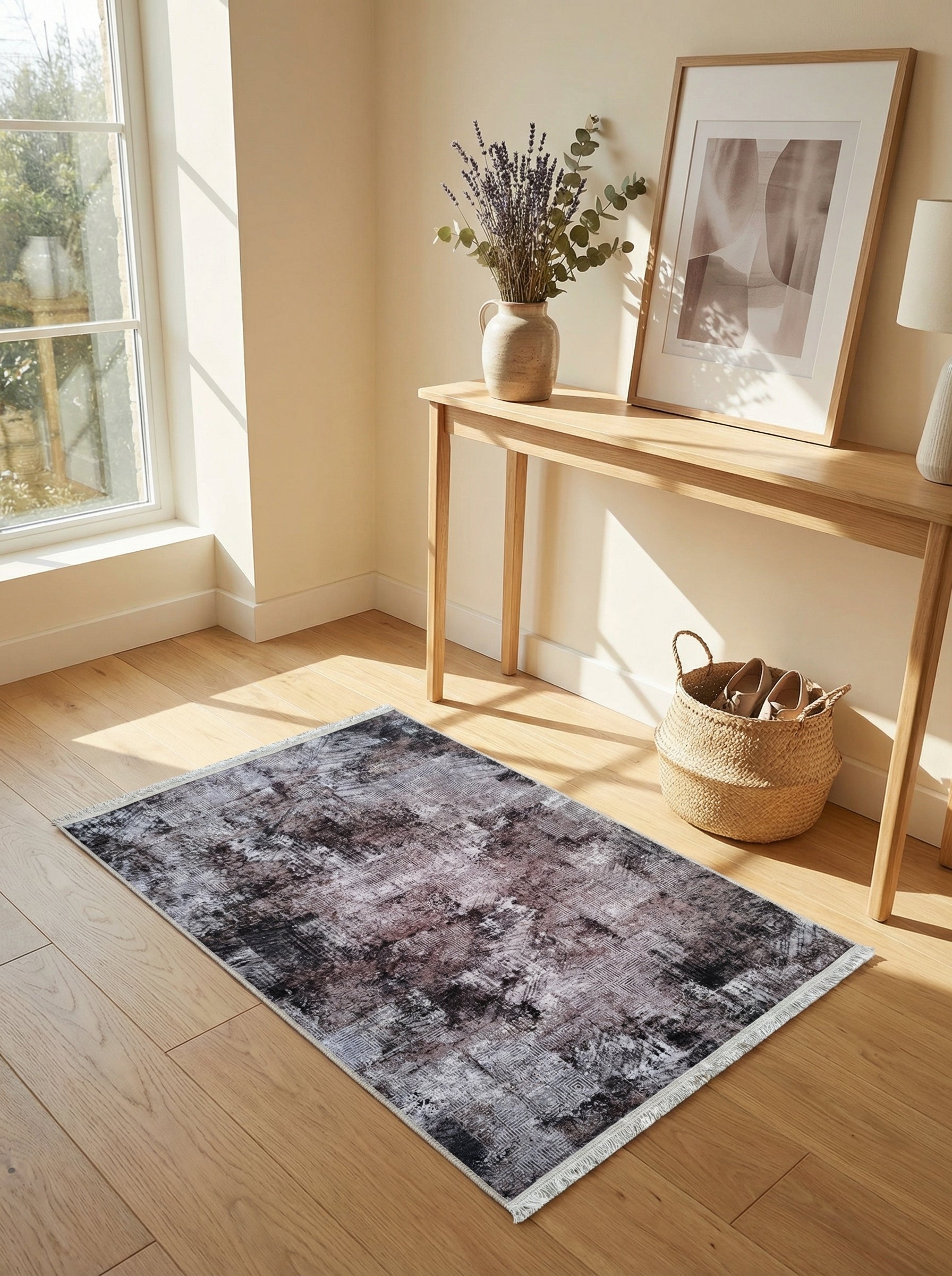Tapis rectangulaire à motifs abstraits gris foncé, noir et blanc cassé avec des franges, posé sur un parquet clair dans une pièce lumineuse