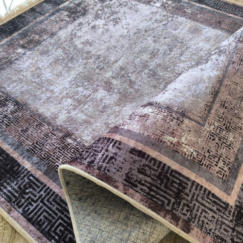 Tapis rectangulaire aux teintes grises et beiges avec un motif abstrait central et des bordures géométriques foncées, partiellement roulé