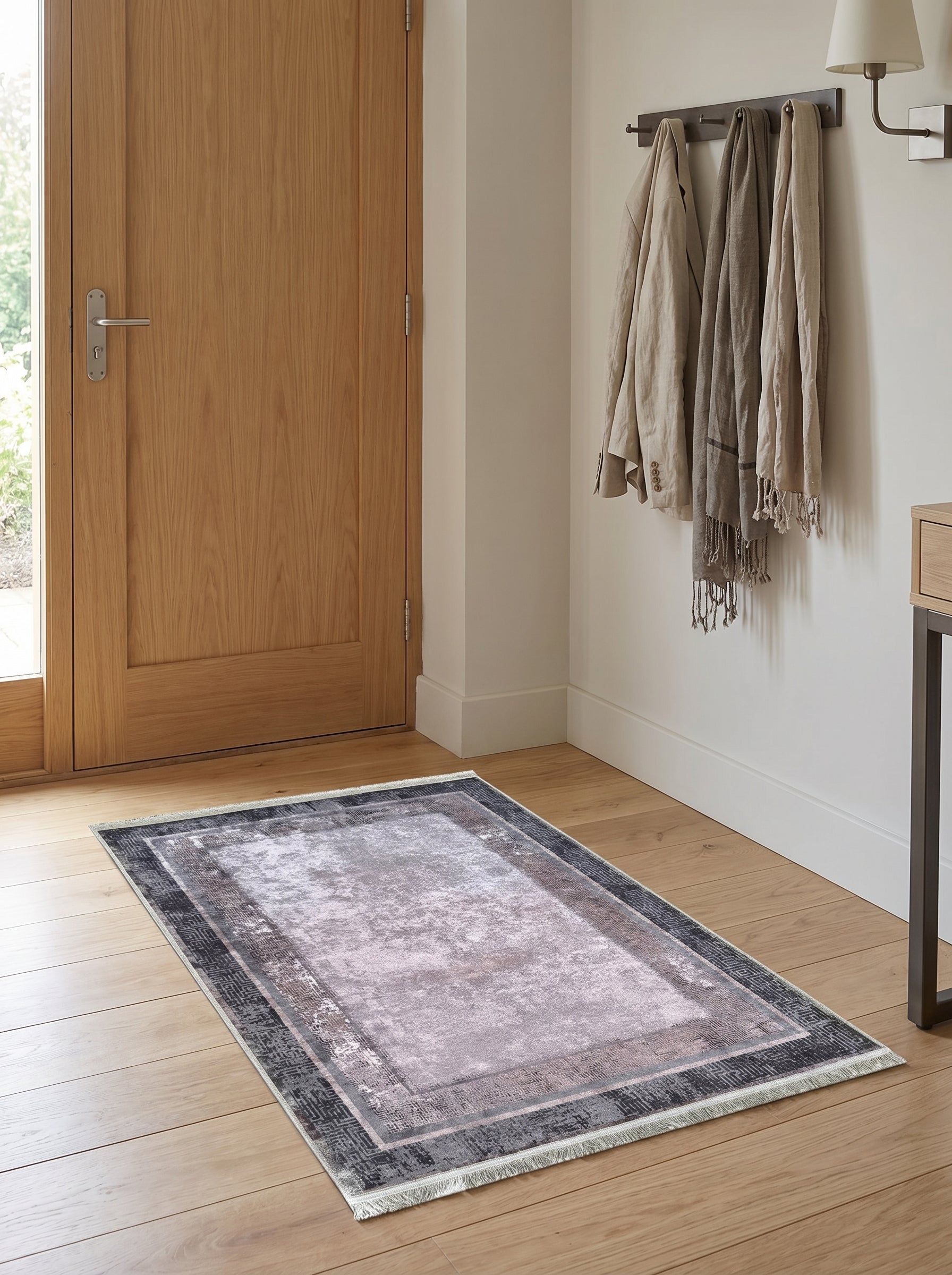 Tapis rectangulaire à motif abstrait gris, mauve et noir avec bordures géométriques, posé sur un parquet clair dans une entrée