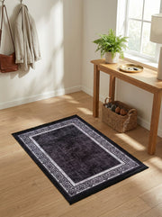 Tapis rectangulaire noir délavé, bordure gris clair à motifs floraux, sur un parquet clair dans une entrée lumineuse