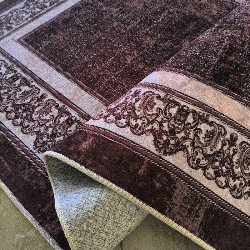 Tapis marron foncé à motifs vieillis et bordures beige clair ornementées de volutes, partiellement roulé