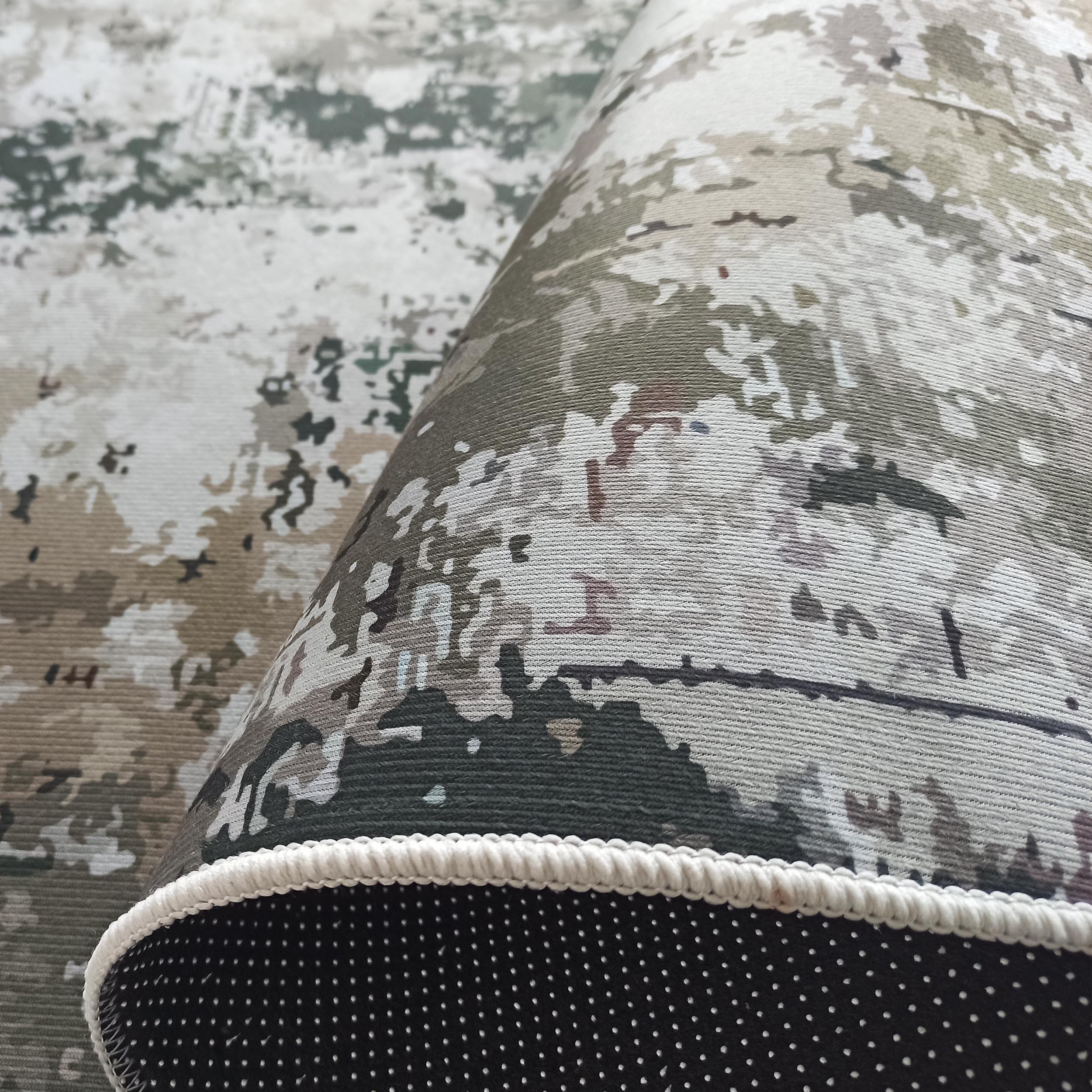 Tapis roulé à motifs abstraits verts, gris, beiges et blancs, avec une bordure tressée blanche et un dessous noir à pois antidérapants