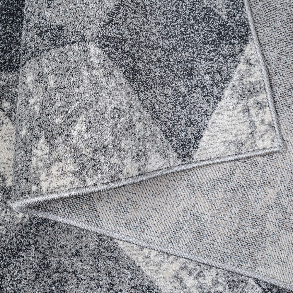Tapis gris à texture mouchetée et motifs abstraits sombres et clairs, avec un coin relevé révélant le revers tissé