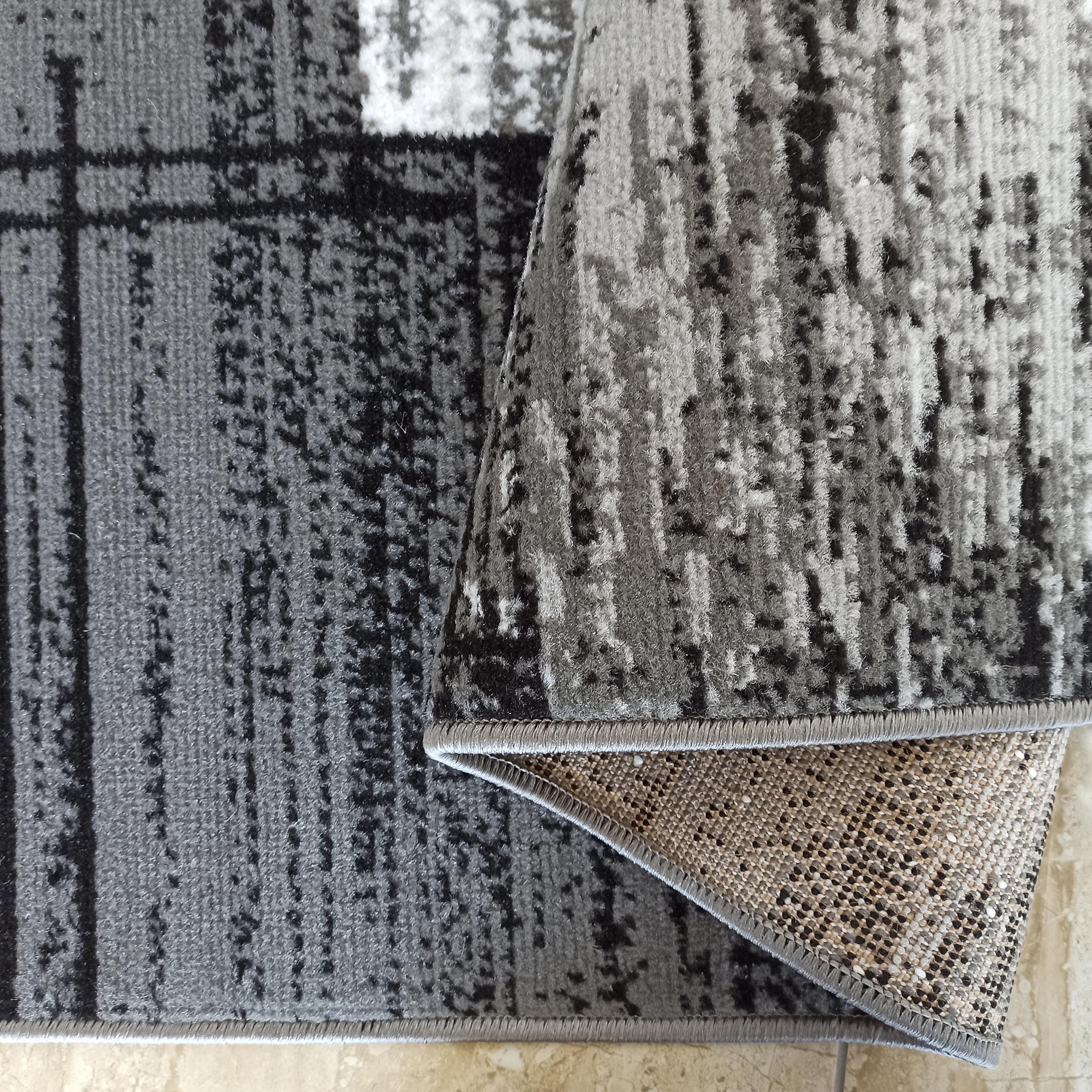 Tapis aux teintes de gris, noir et blanc avec un motif abstrait. Un coin est replié, révélant son envers tissé moucheté et sa bordure ourlée