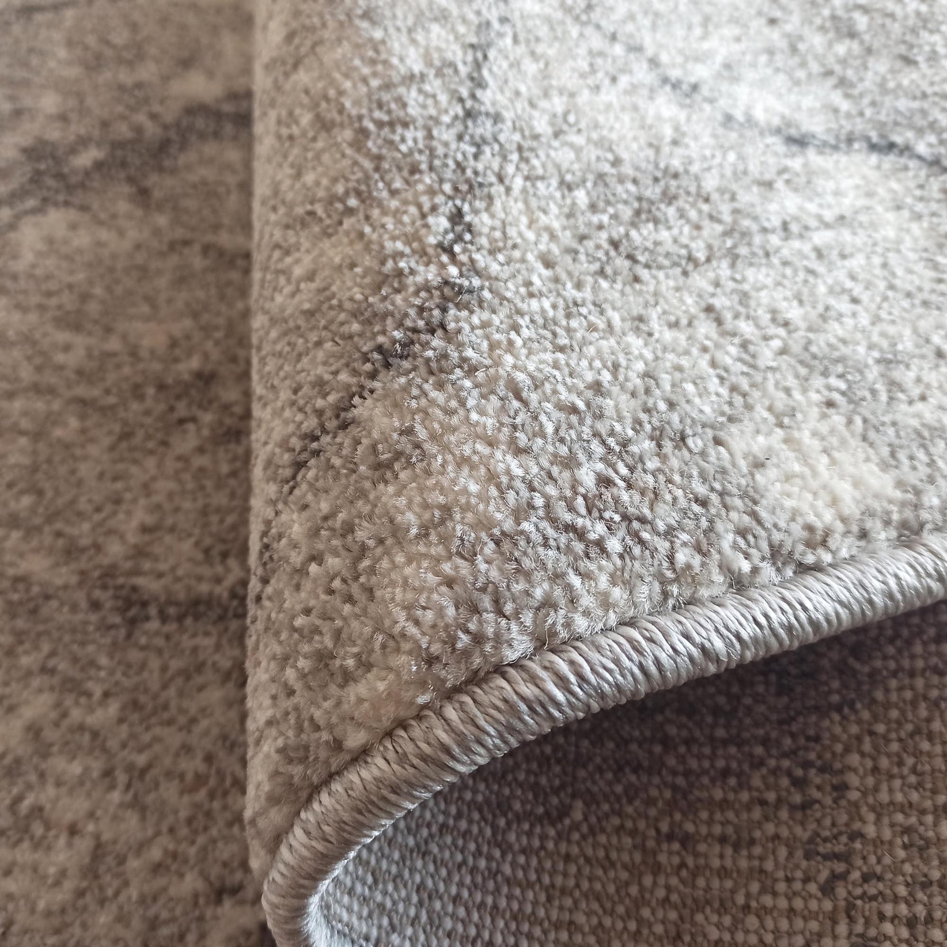 Tapis doux et texturé, en gros plan, présentant des tons de gris, blanc et beige. Son motif est abstrait marbré et sa bordure est ourlée