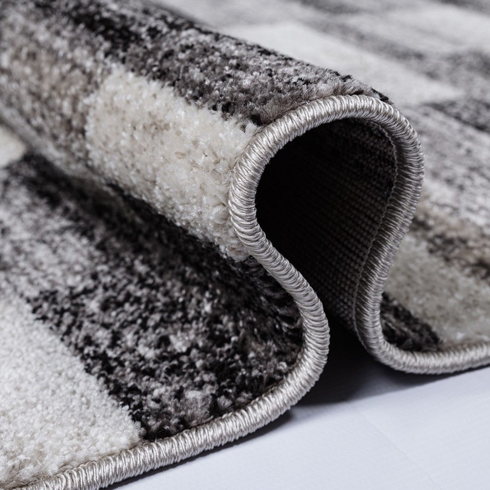 Gros plan sur un tapis texturé gris, noir et blanc, avec un motif abstrait, partiellement enroulé montrant sa bordure cousue