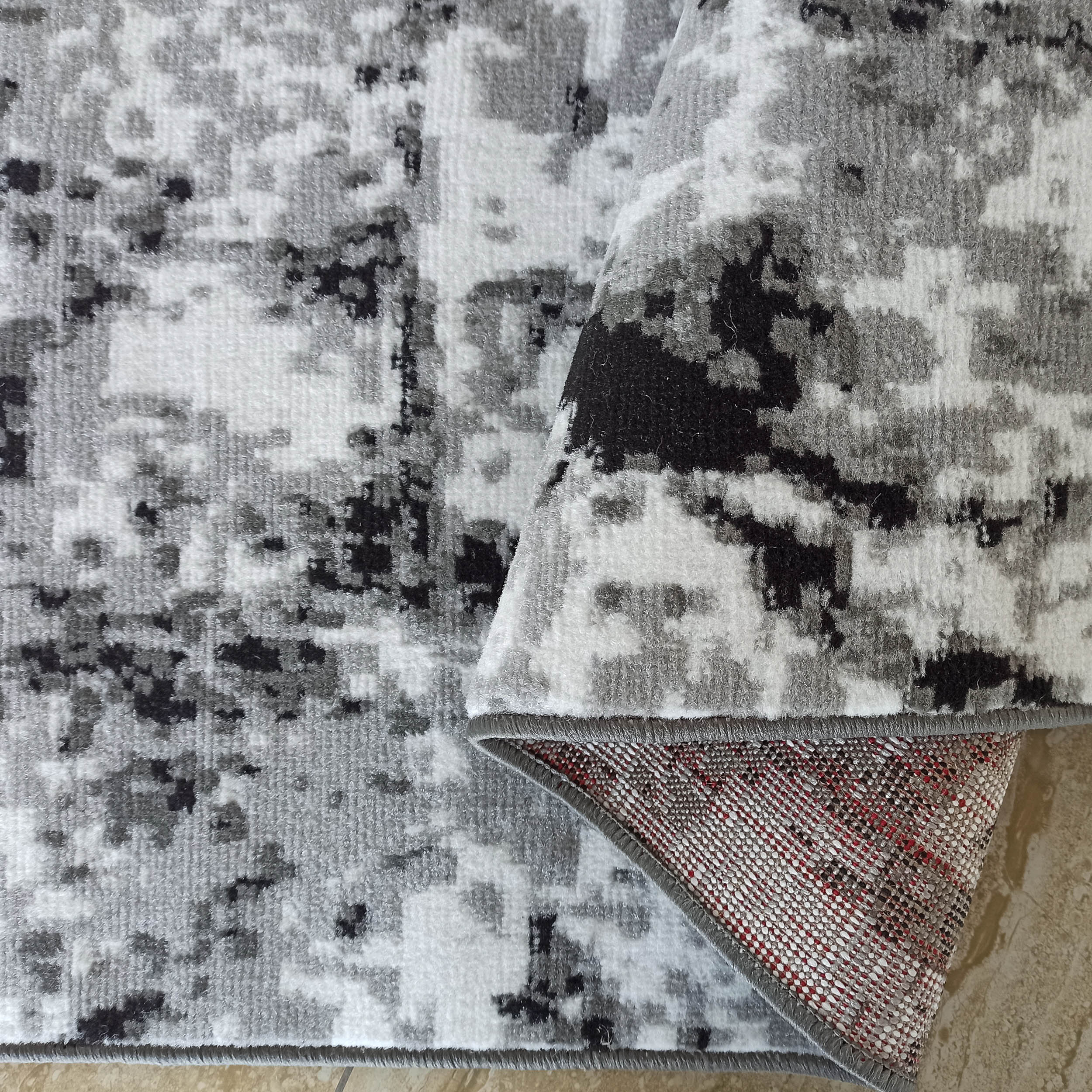 Tapis moelleux aux motifs abstraits gris, noir et blanc, partiellement enroulé pour révéler son envers tissé