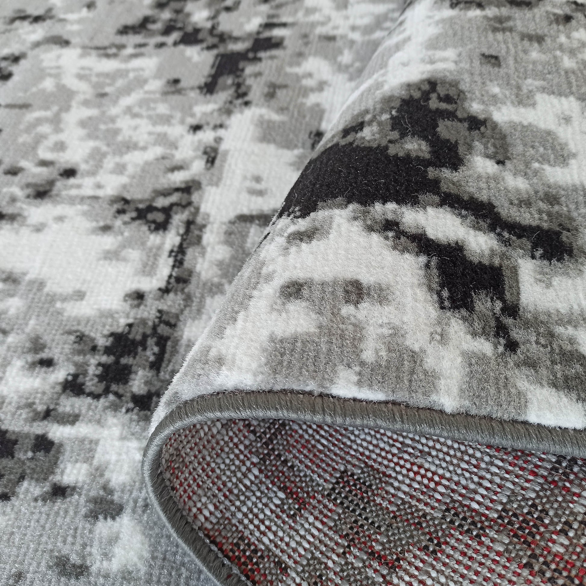 Tapis à poils courts gris, blanc et noir avec un motif abstrait, dont un coin est relevé, dévoilant le dessous tissé
