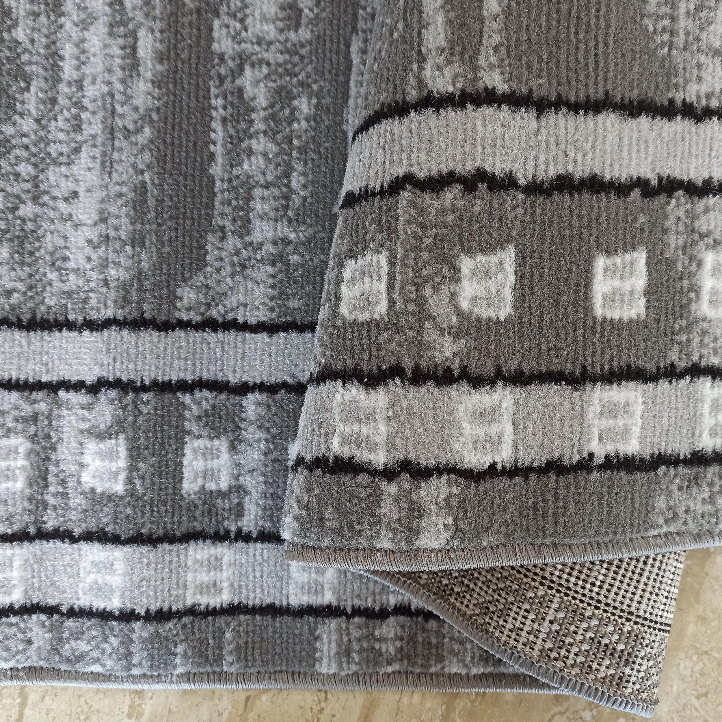 Tapis gris et blanc à motifs abstraits, avec des bandes horizontales noires et blanches contenant des carrés. Une partie du tapis est pliée, révélant son envers tissé