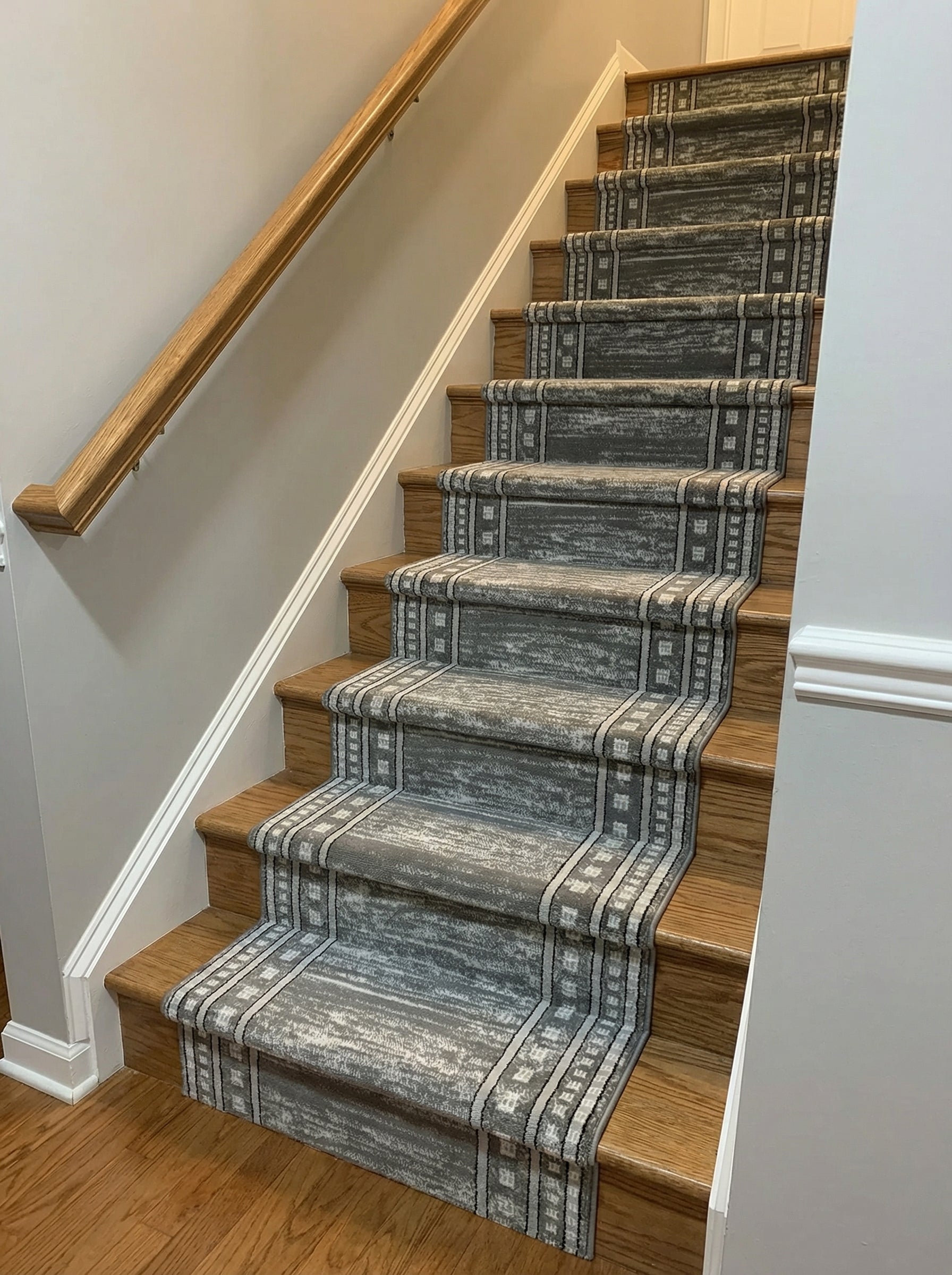 Tapis d'escalier gris et blanc à l'aspect vieilli avec des bordures géométriques sur des marches en bois clair dans une entrée