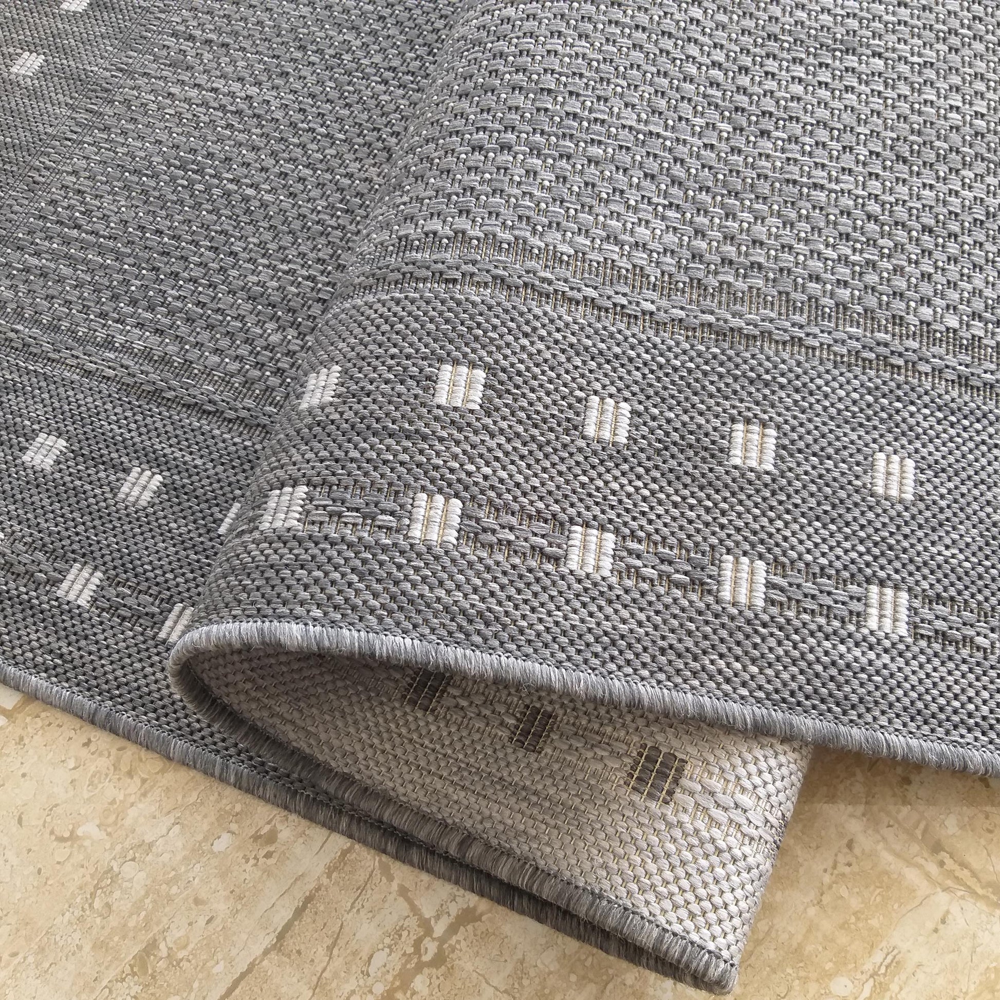 Tapis gris à tissage serré avec des motifs en barres blanches. Il est partiellement roulé sur un sol carrelé beige