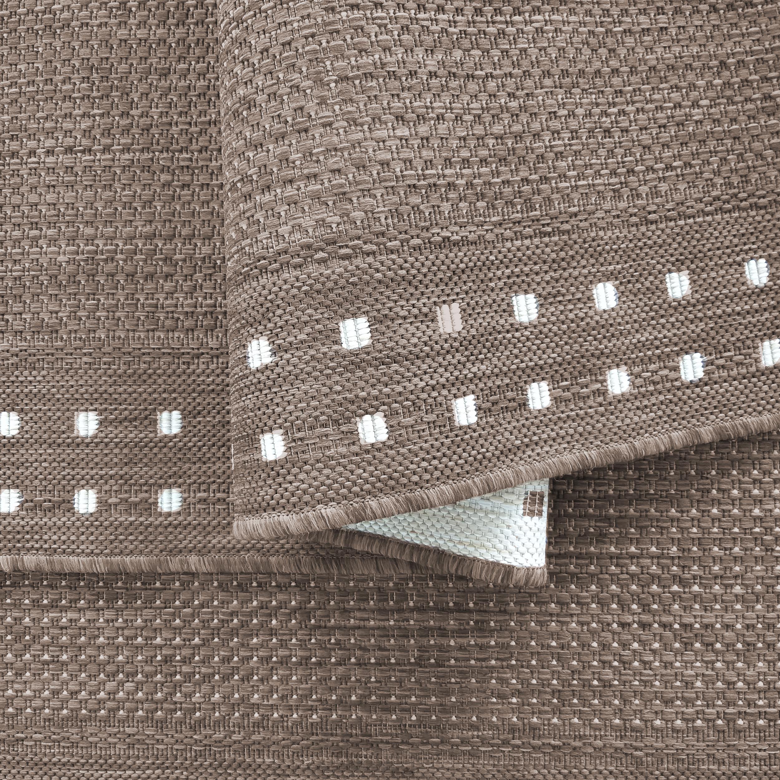 Tapis tissé marron-taupe plié montrant une bordure à motifs de carrés blancs et des franges, l'envers clair est visible