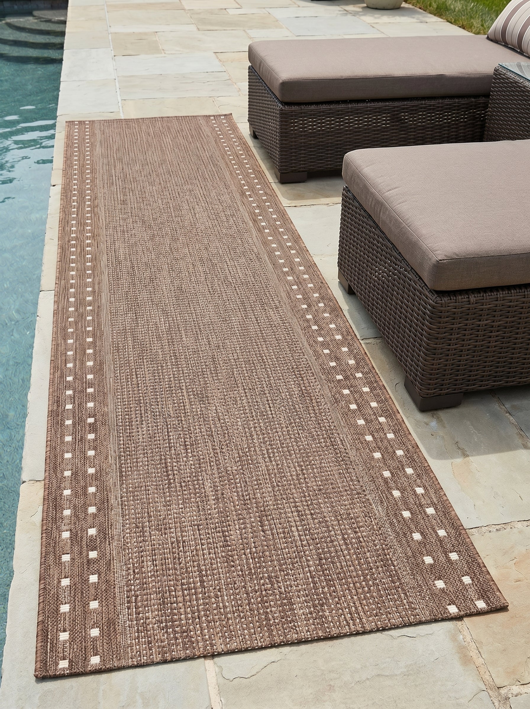 Tapis rectangulaire marron tissé avec des motifs de carrés crème le long des bords, sur une terrasse près d'une piscine