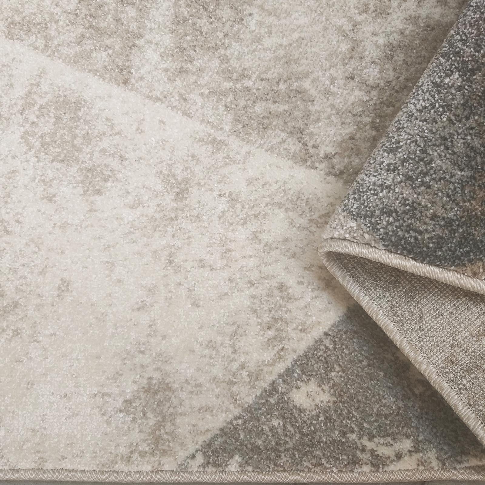 Tapis à motifs abstraits beige et gris, partiellement plié, montrant sa texture douce et son envers tissé
