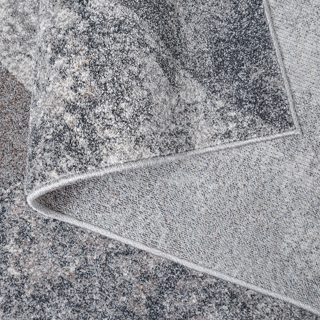 Tapis à la surface mouchetée et abstraite, mélange de gris, blanc et quelques touches de beige, un coin relevé dévoilant son envers tissé