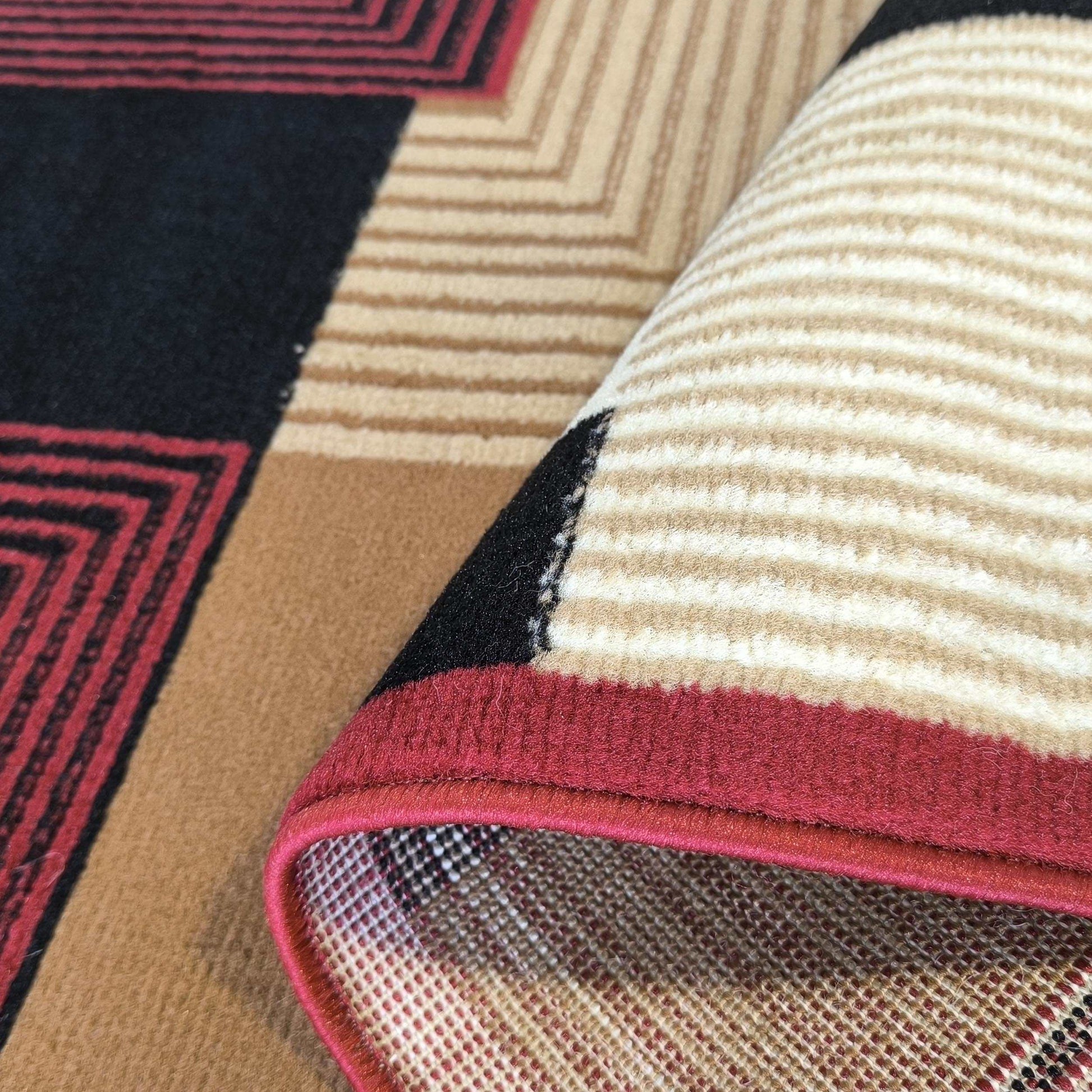 Tapis à poils courts aux motifs géométriques rouge, noir, beige et marron. Un pan est roulé, révélant la doublure maillée et la bordure rouge