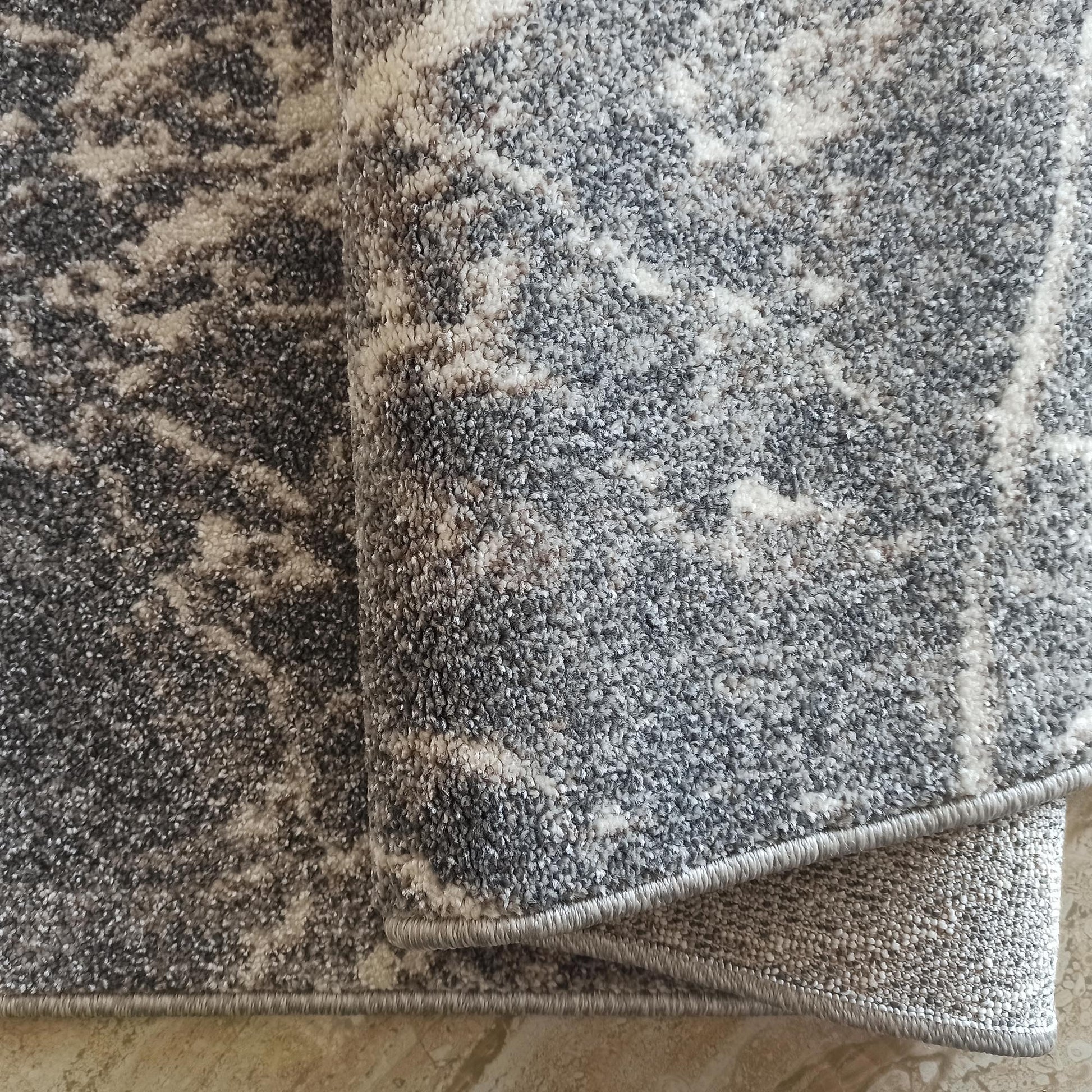 Tapis moelleux gris et blanc cassé à motif abstrait irrégulier, avec une bordure surpiquée