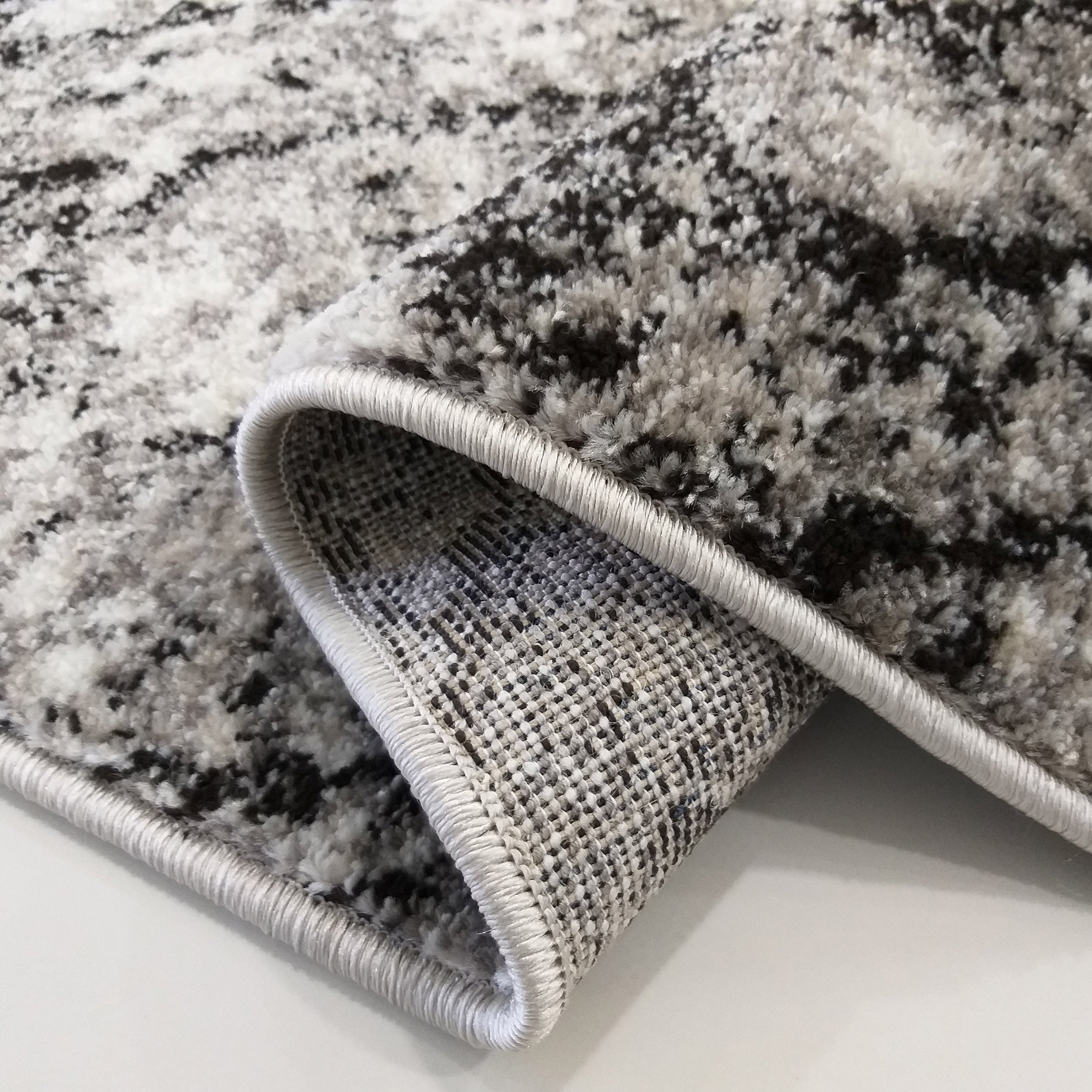 Tapis moelleux aux motifs abstraits gris, blancs et noirs, avec une bordure claire et le dos tissé visible