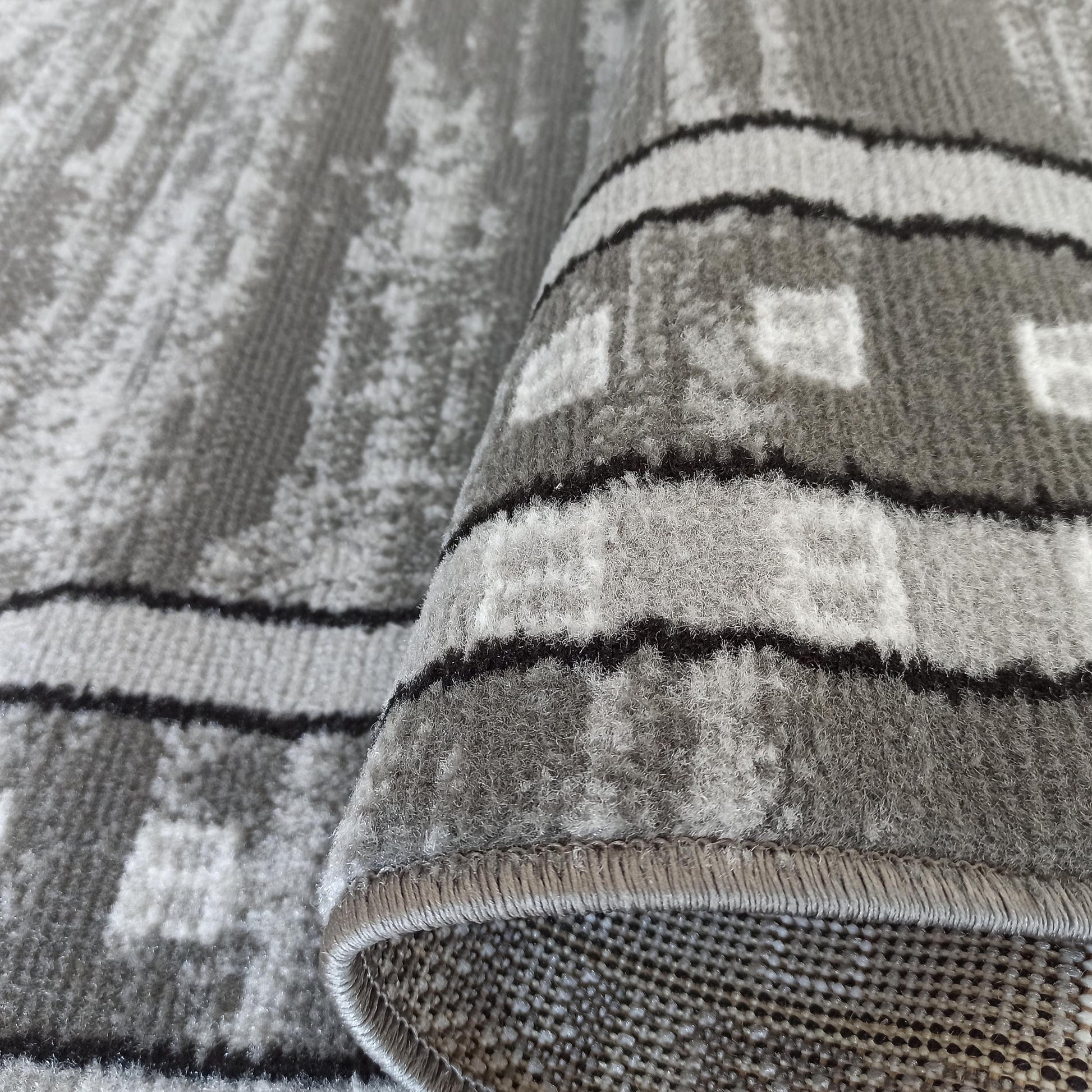 Tapis aux motifs gris, noirs et blancs. Un bord est replié, montrant la texture de ses poils denses et son envers tissé