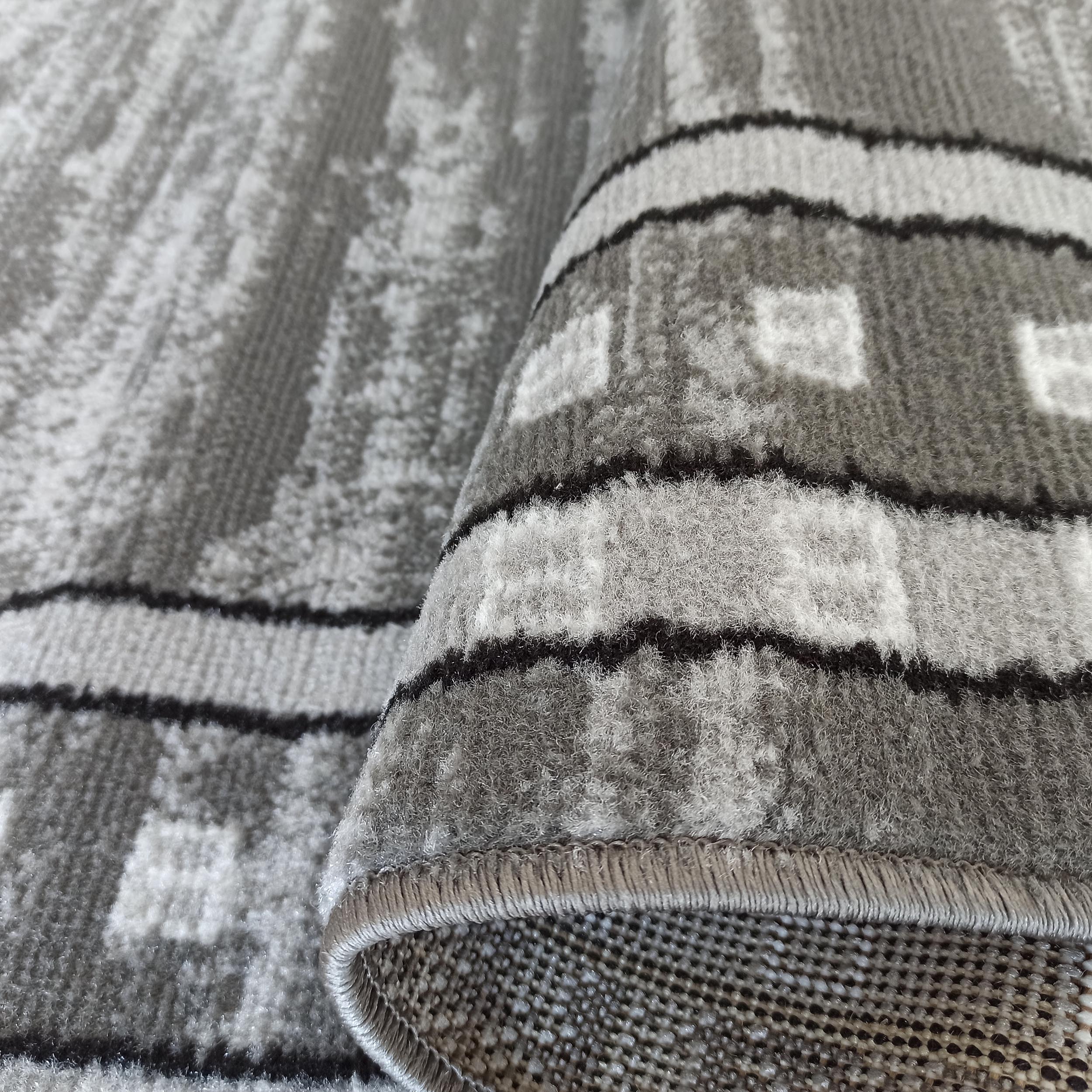 Tapis aux motifs gris, noirs et blancs. Un bord est replié, montrant la texture de ses poils denses et son envers tissé