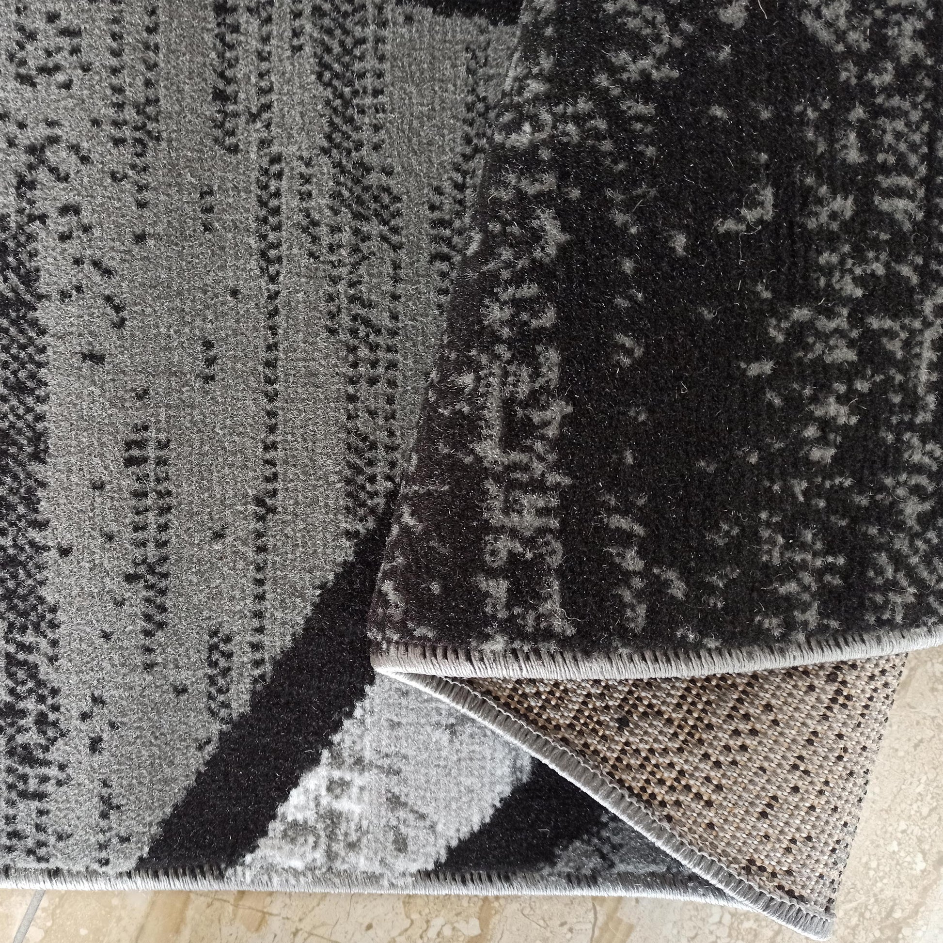 Tapis doux gris et noir aux motifs abstraits mouchetés Un coin plié révèle l'envers tissé beige clair à pois foncés