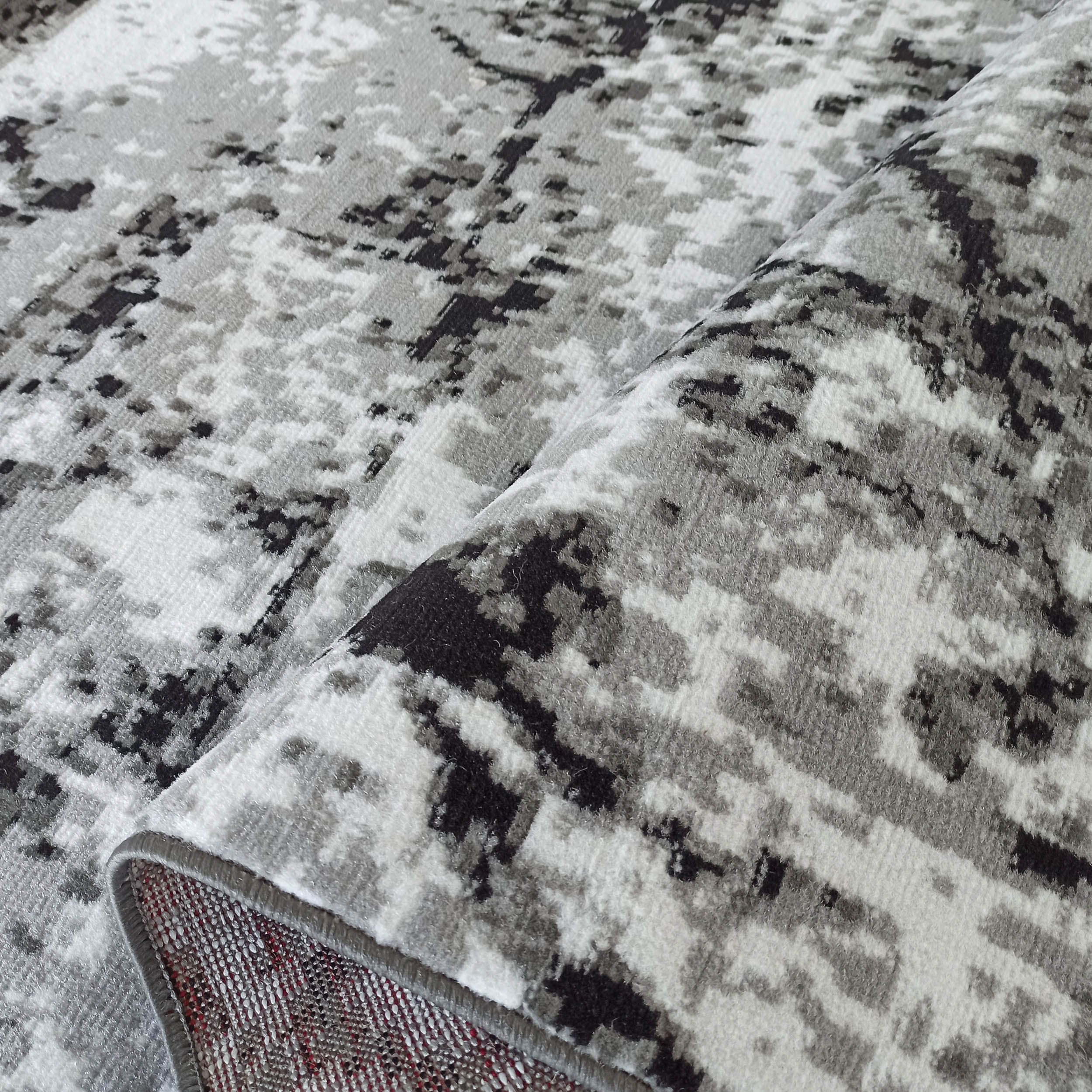 Tapis aux motifs abstraits en dégradé de gris, noir et blanc, présentant une texture douce. Il est partiellement plié, montrant son envers