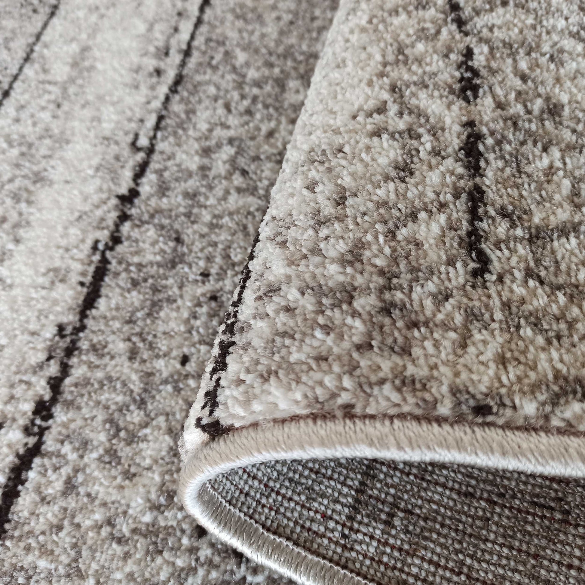 Gros plan d'un tapis à poils longs aux teintes de beige et crème, avec des motifs de rayures sombres. Un coin du tapis est relevé, révélant son envers