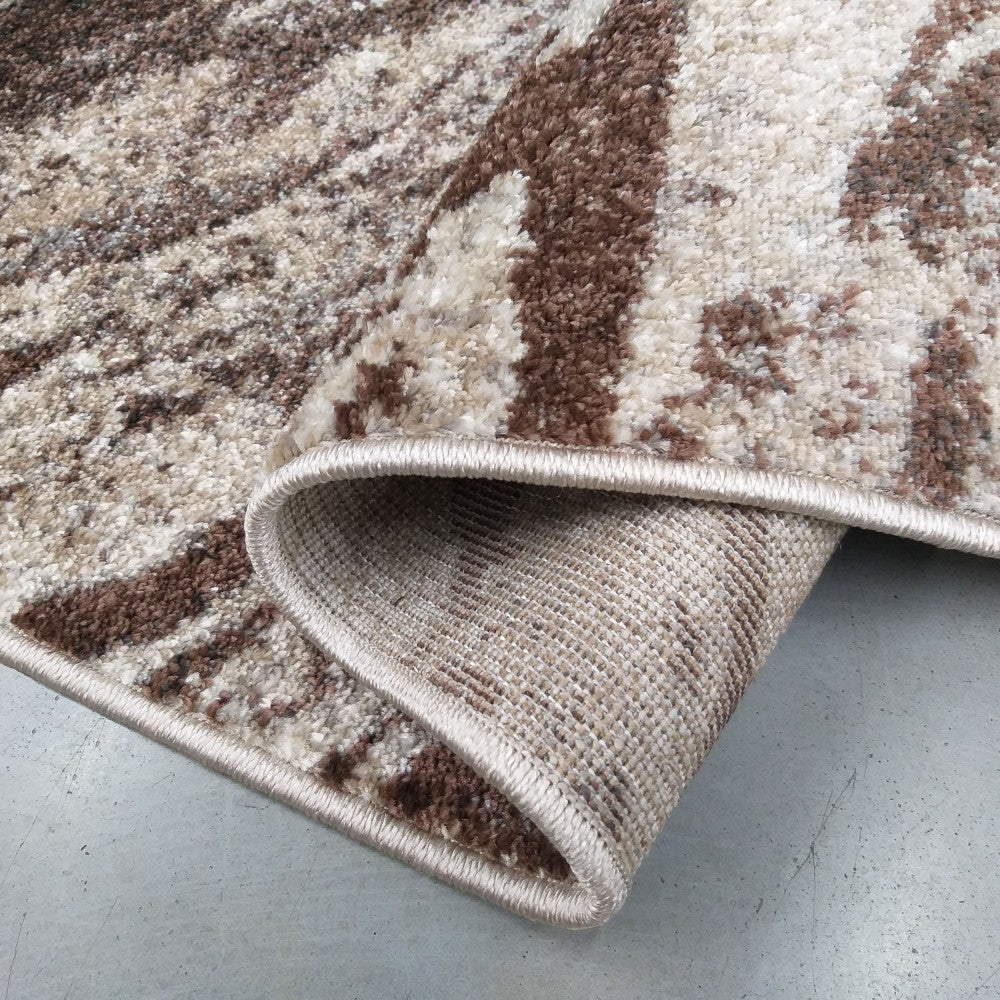 Tapis moelleux à motifs abstraits marron et beige, partiellement roulé sur un sol gris, montrant son envers tissé et sa bordure claire