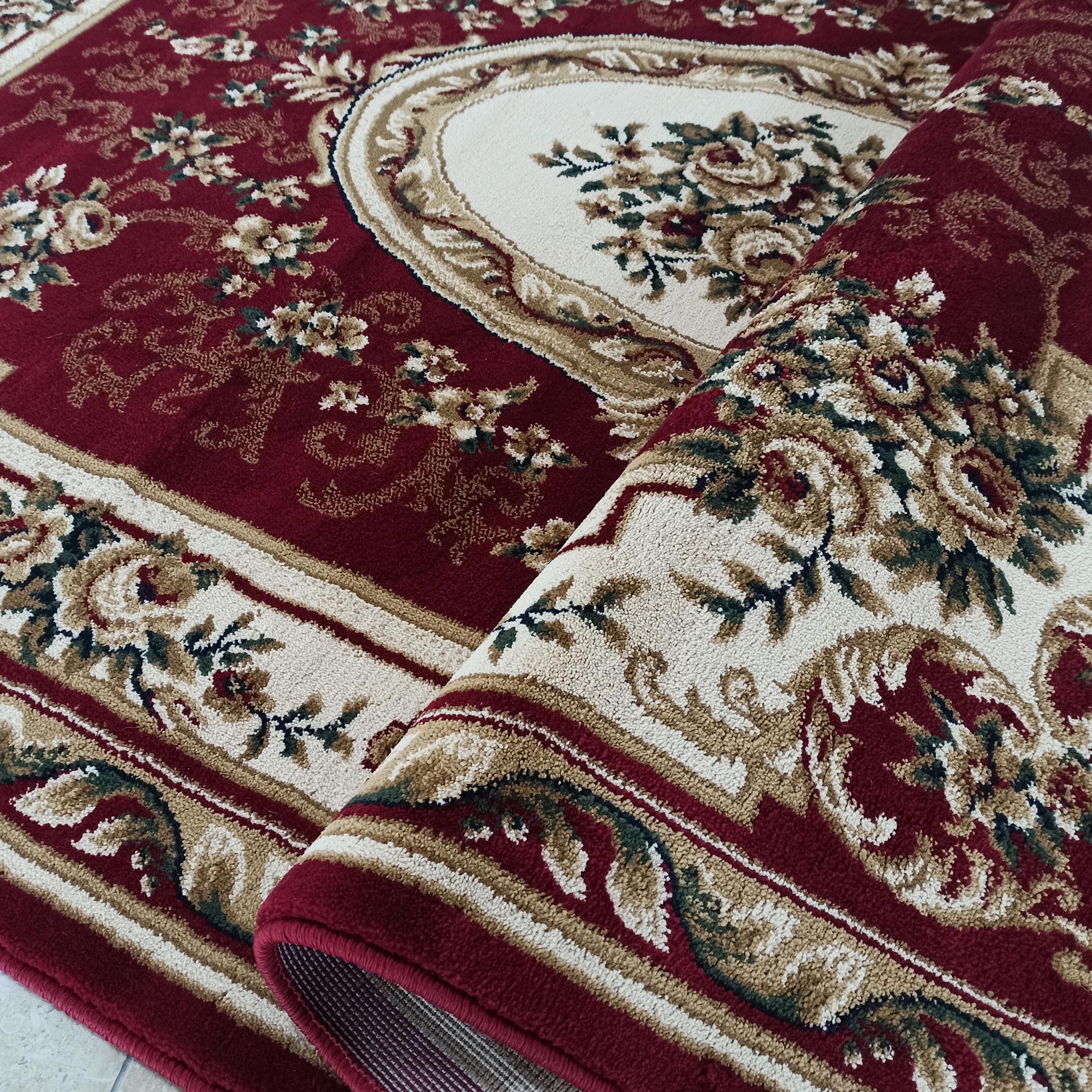 Tapis bordeaux avec un grand motif central ovale crème. Des détails floraux verts et beiges ornent le tapis, visiblement roulé