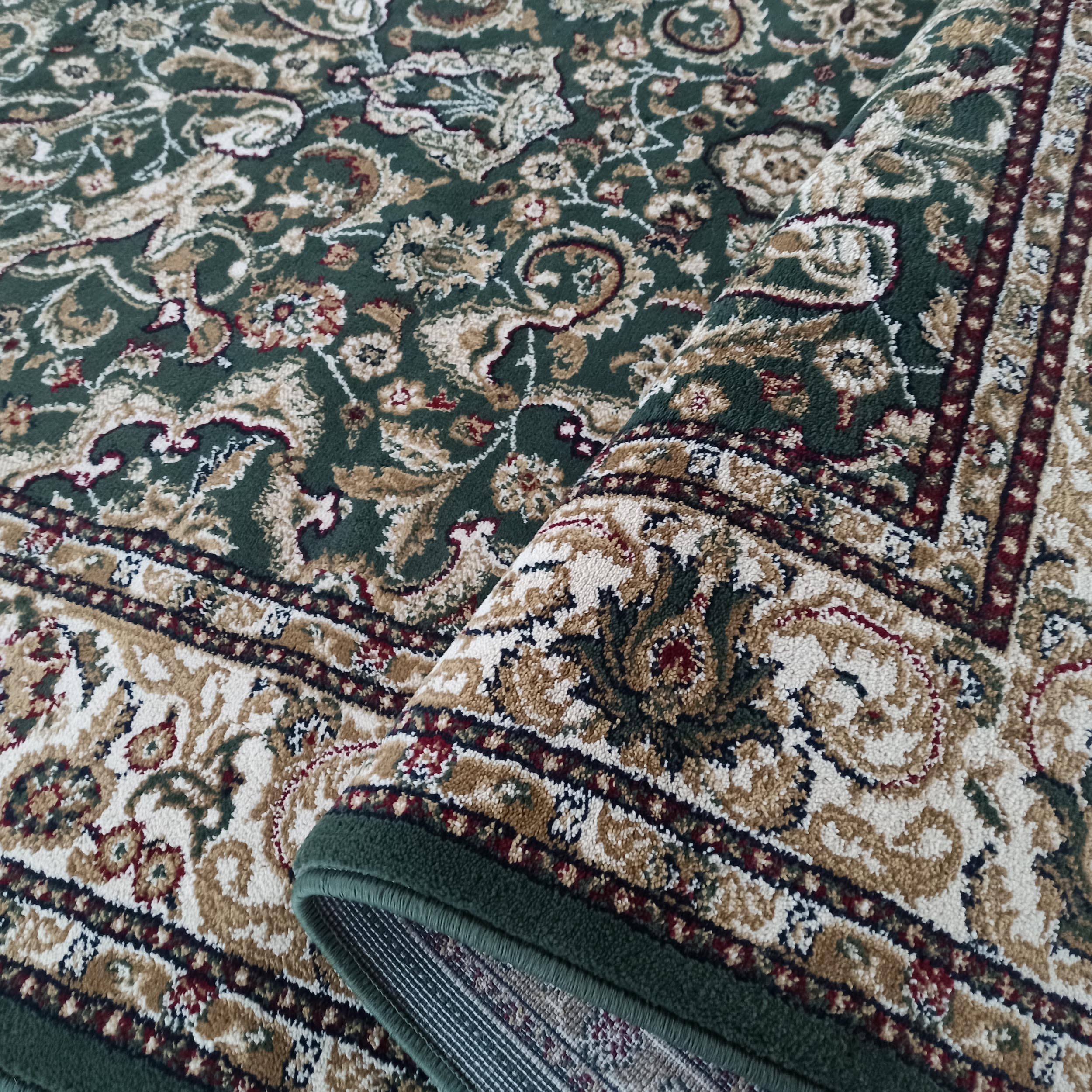 Tapis vert foncé avec motifs floraux et cachemire beiges, crème et rouge bordeaux, partiellement roulé