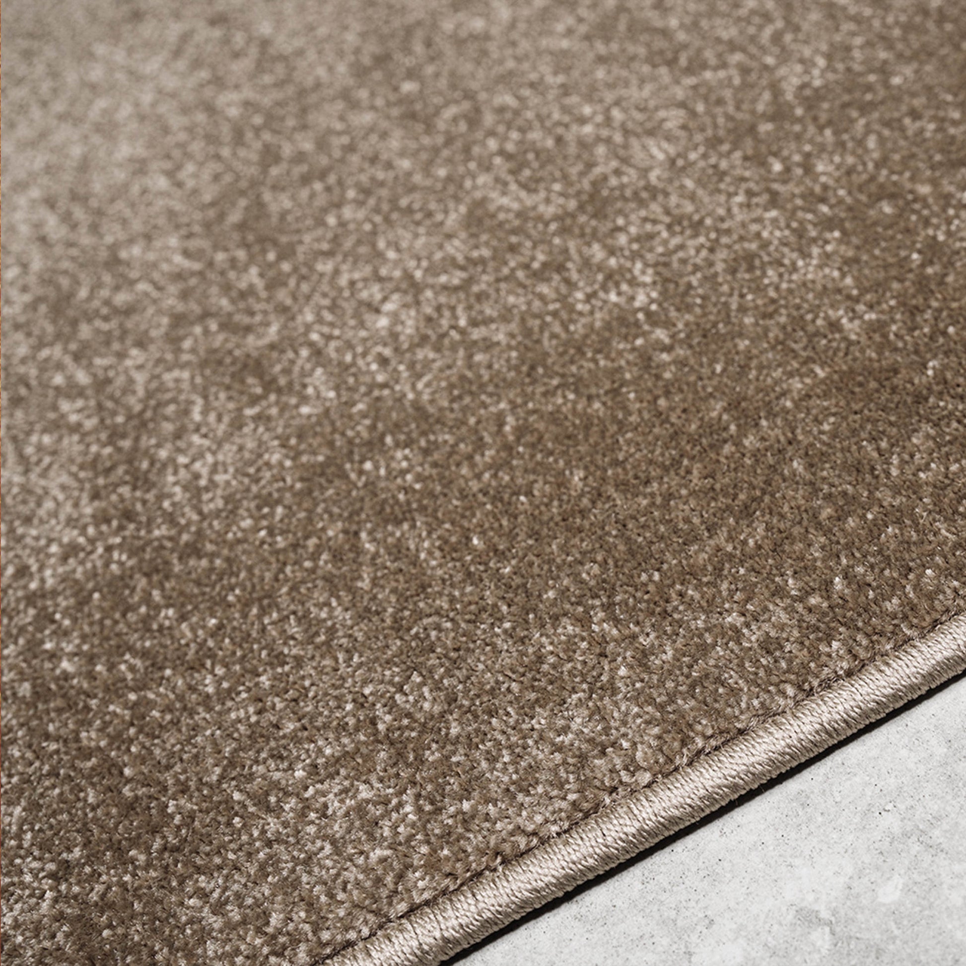 Tapis doux de couleur beige-brun, avec une texture mouchetée de nuances claires et sombres. Son bord est visible sur un sol clair
