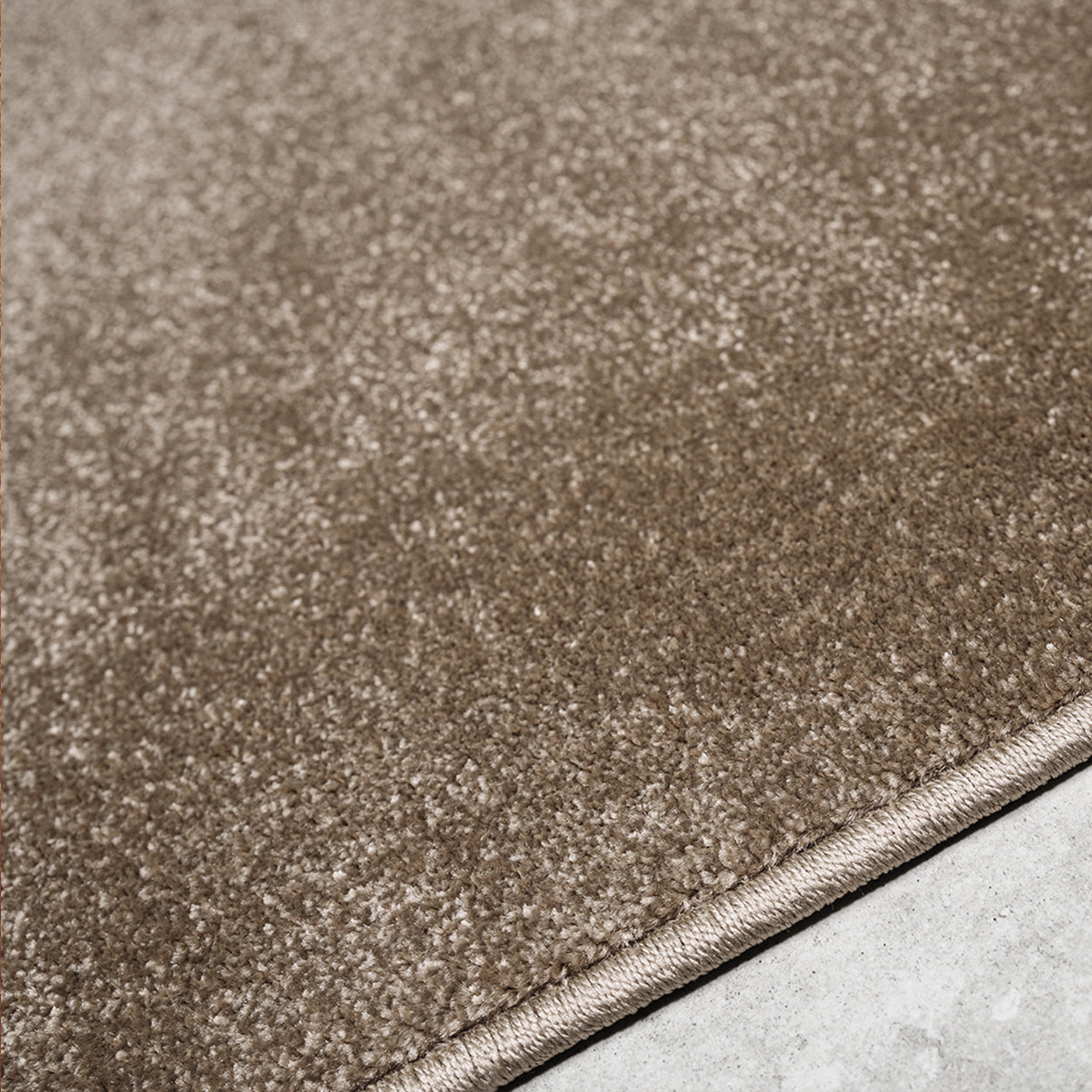 Tapis doux de couleur beige-brun, avec une texture mouchetée de nuances claires et sombres. Son bord est visible sur un sol clair