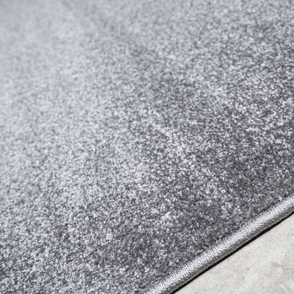 Un tapis gris clair à poils doux et brillants, avec une bordure cousue visible sur un sol gris