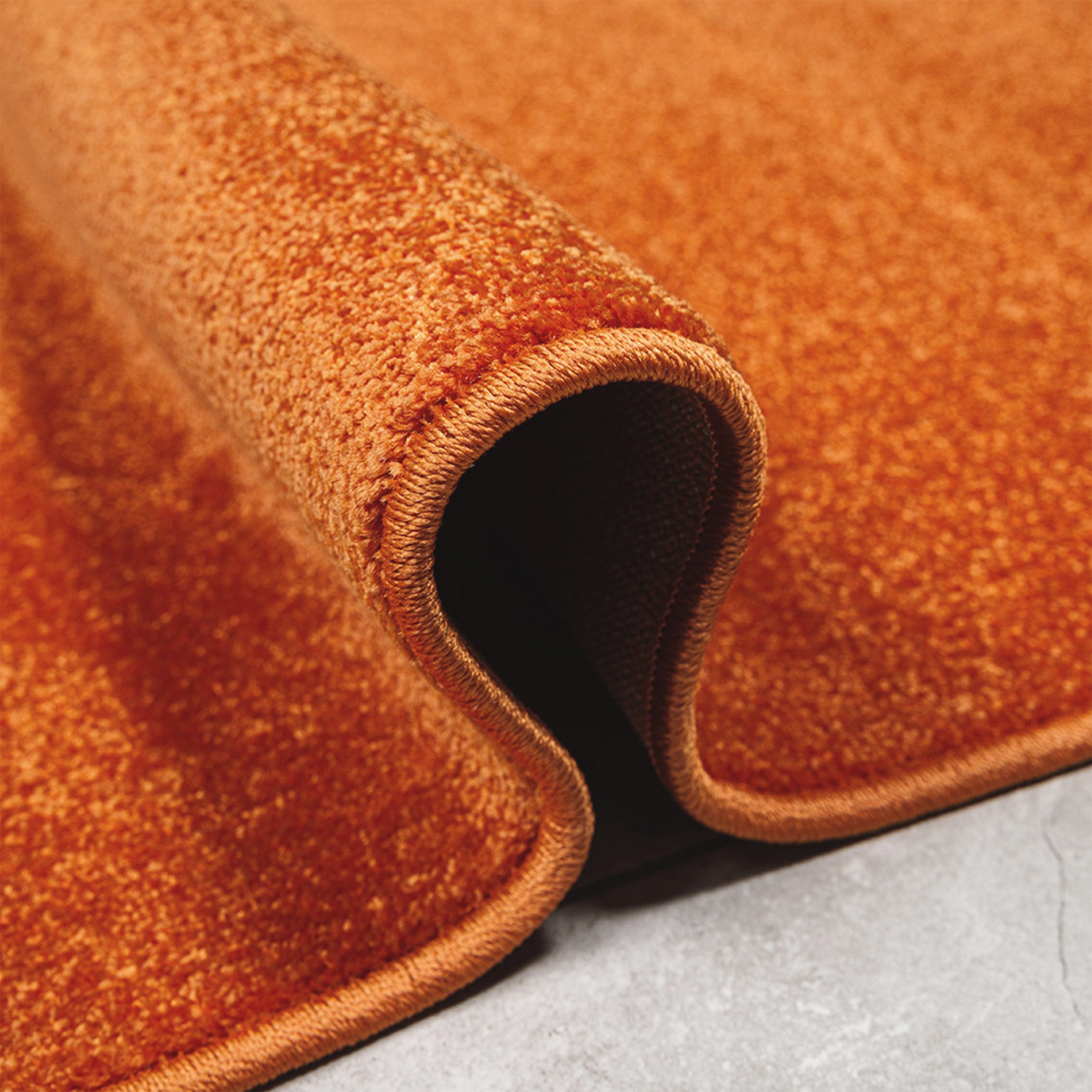Tapis orange rouille à poils courts et dense, partiellement roulé, avec un bord surpiqué, sur un sol gris clair