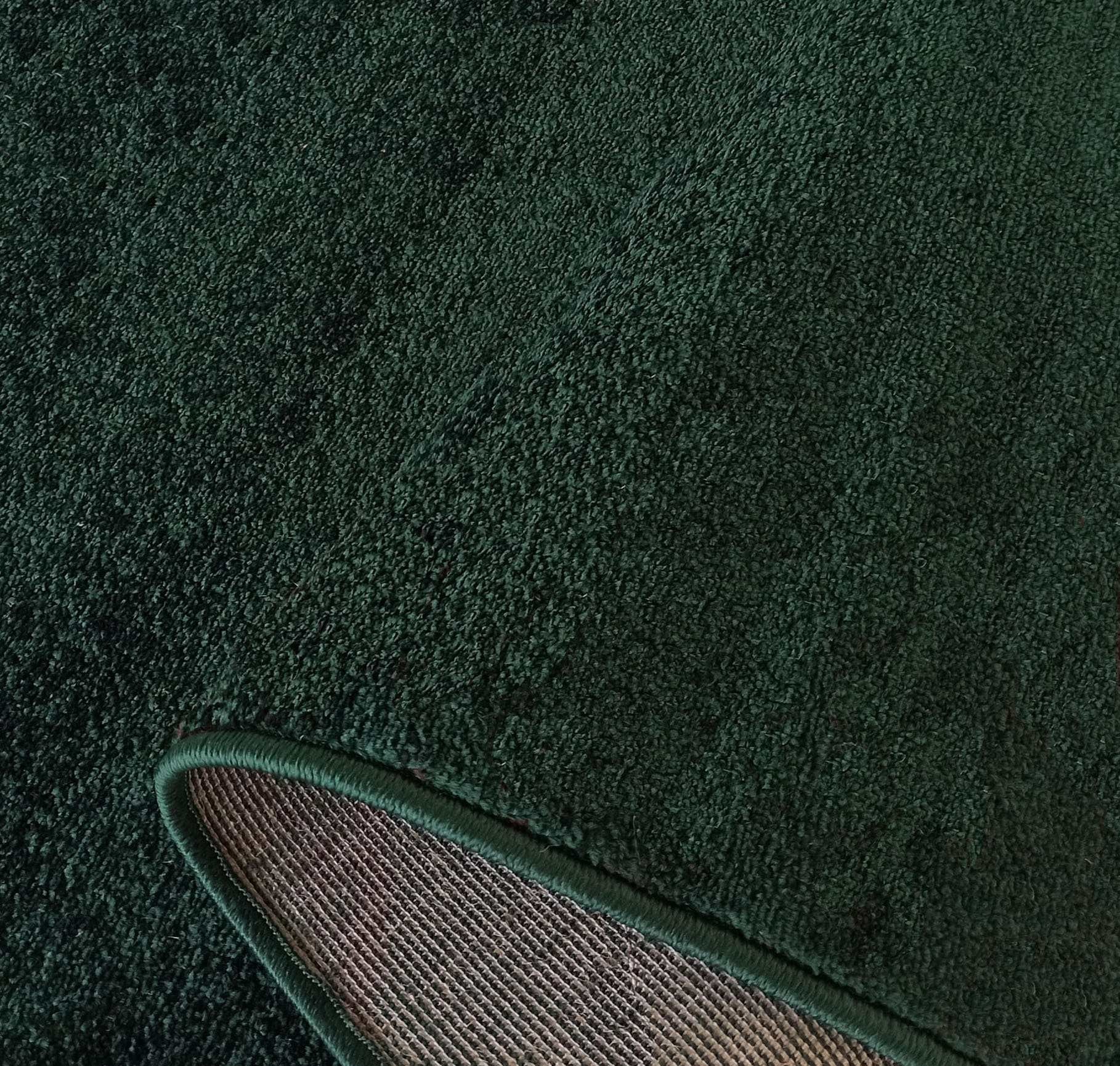 Tapis vert foncé en velours bouclé doux, avec un coin relevé montrant son dos tissé marron et son ourlet vert
