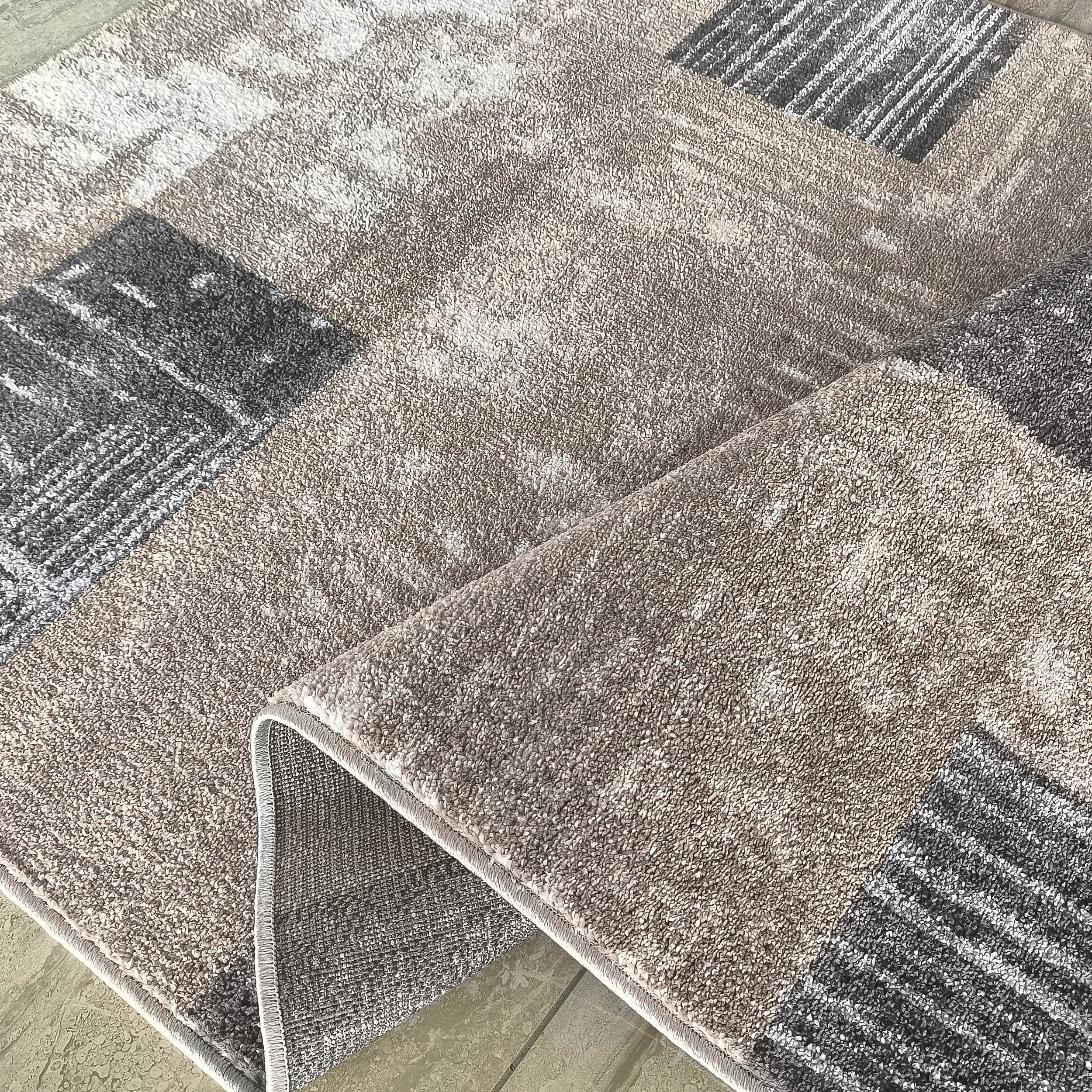 Tapis moelleux aux motifs abstraits beiges, gris foncés et blancs, plié sur un carrelage clair, montrant son envers tissé