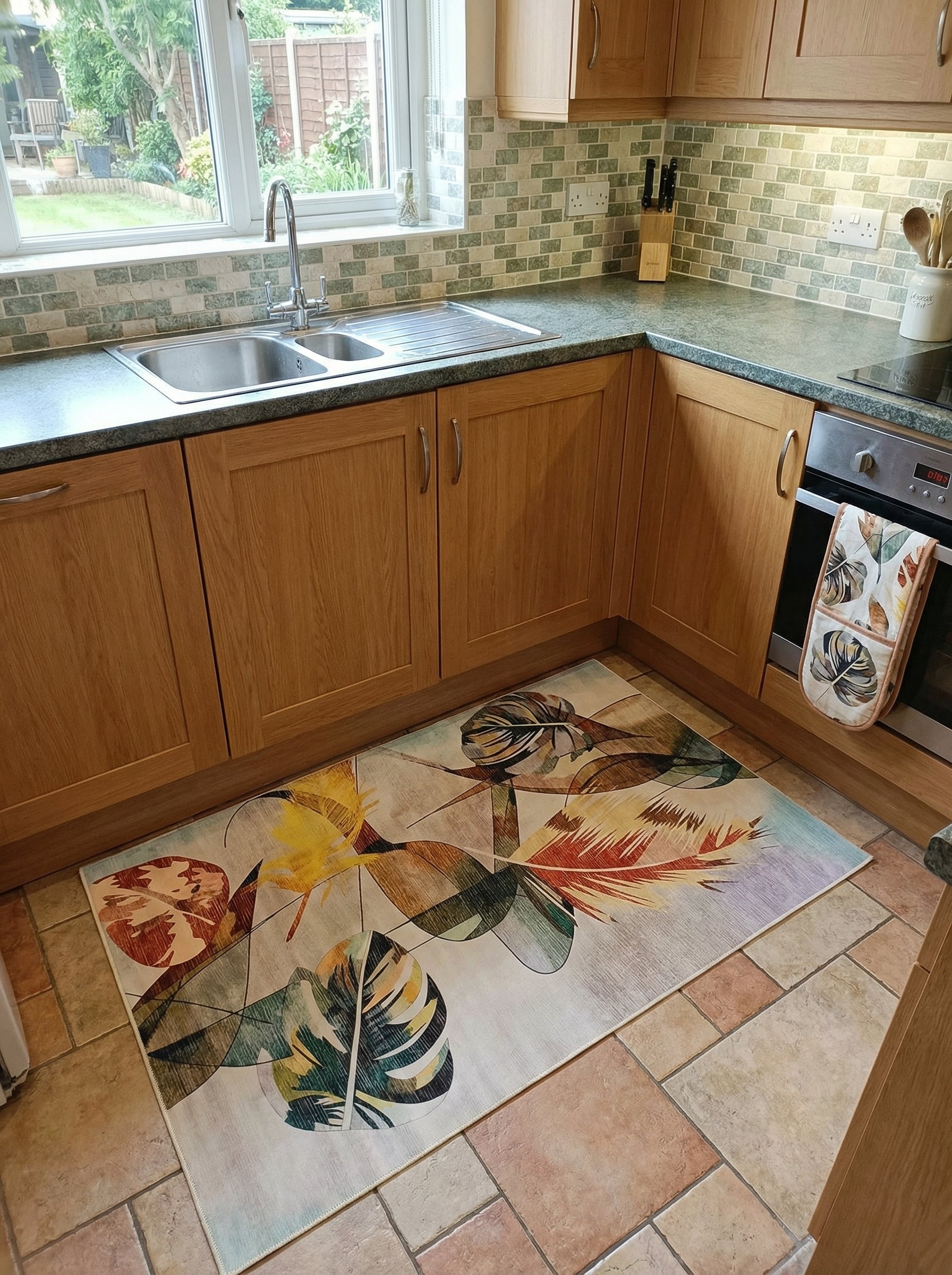 Tapis rectangulaire à motifs de feuilles tropicales stylisées aux couleurs vives (vert, jaune, orange, brun) sur fond clair, dans une cuisine