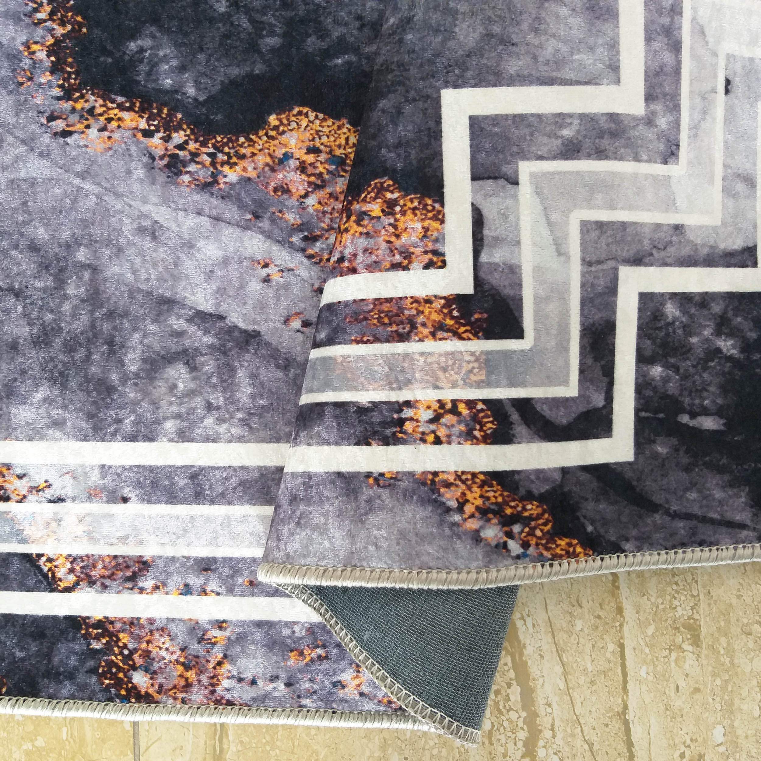 Tapis velouté gris foncé et clair, aux motifs abstraits orange doré et lignes géométriques blanches, partiellement roulé sur un sol carrelé beige, montrant son envers