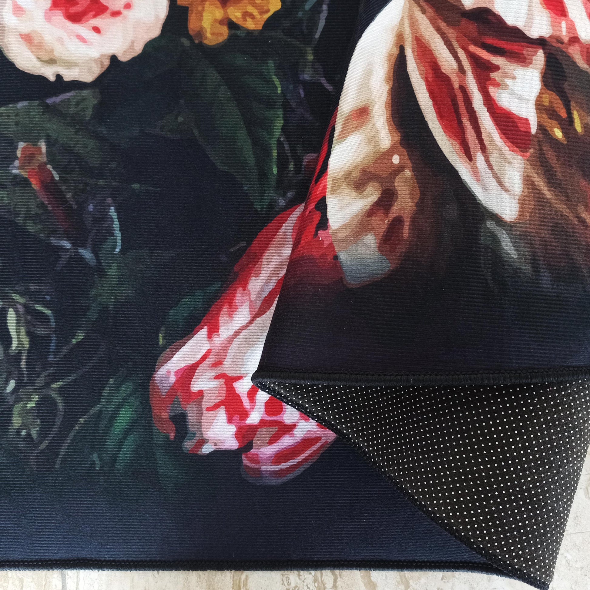 Tapis à motifs floraux détaillés de roses rouges, blanches et orange sur fond bleu foncé, son envers antidérapant à pois blancs est visible