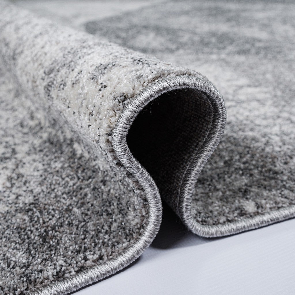 Un tapis gris clair et foncé avec un motif abstrait moucheté, partiellement enroulé et montrant sa bordure soignée